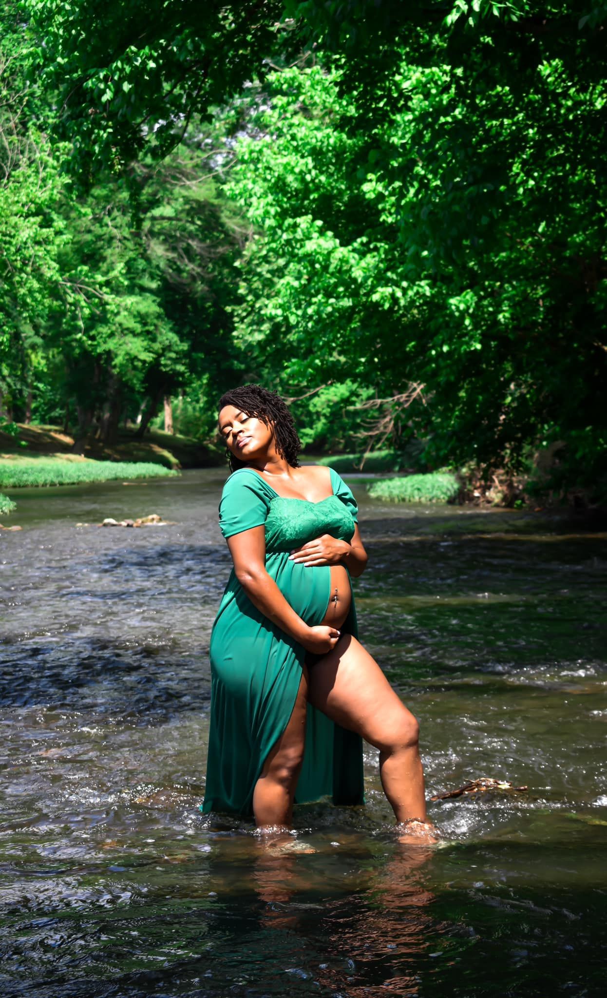 Pregnant woman in flowing teal gown standing in peaceful forest stream surrounded by lush green trees.