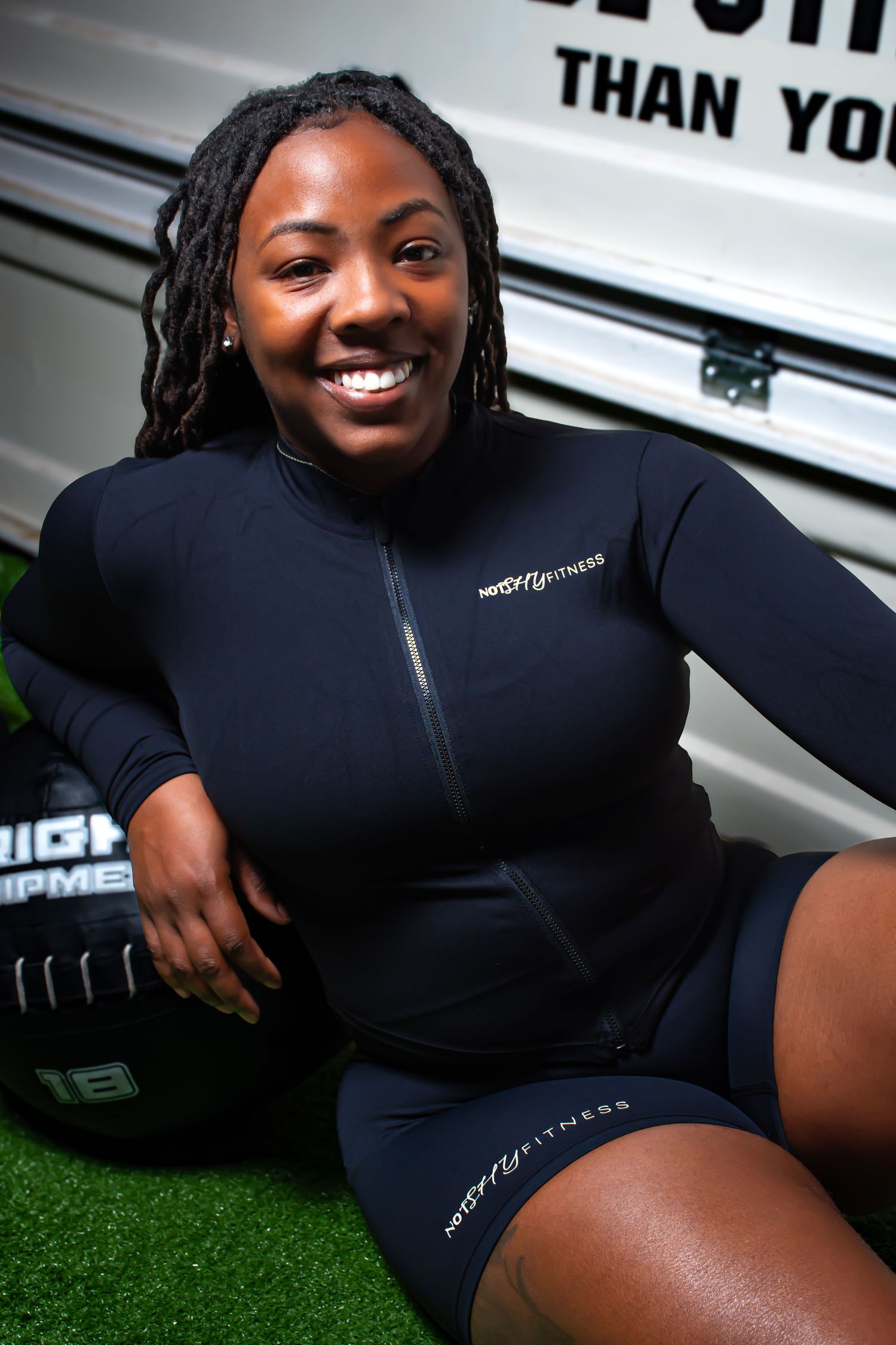 Smiling woman in black VersaFit Fitness athletic outfit sitting on turf near gym equipment outdoors.
