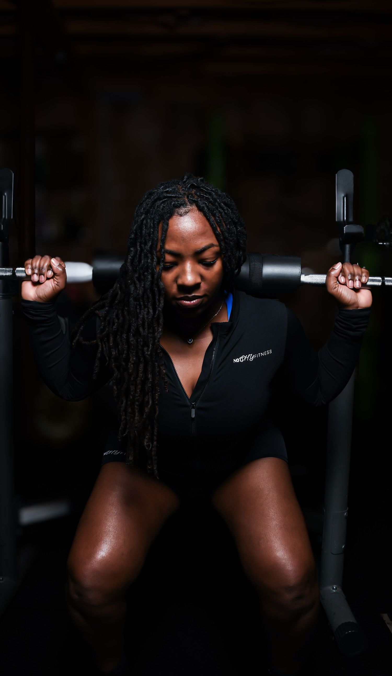A Black woman in a black zip-up top performs a barbell squat exercise at the gym with focused determination.