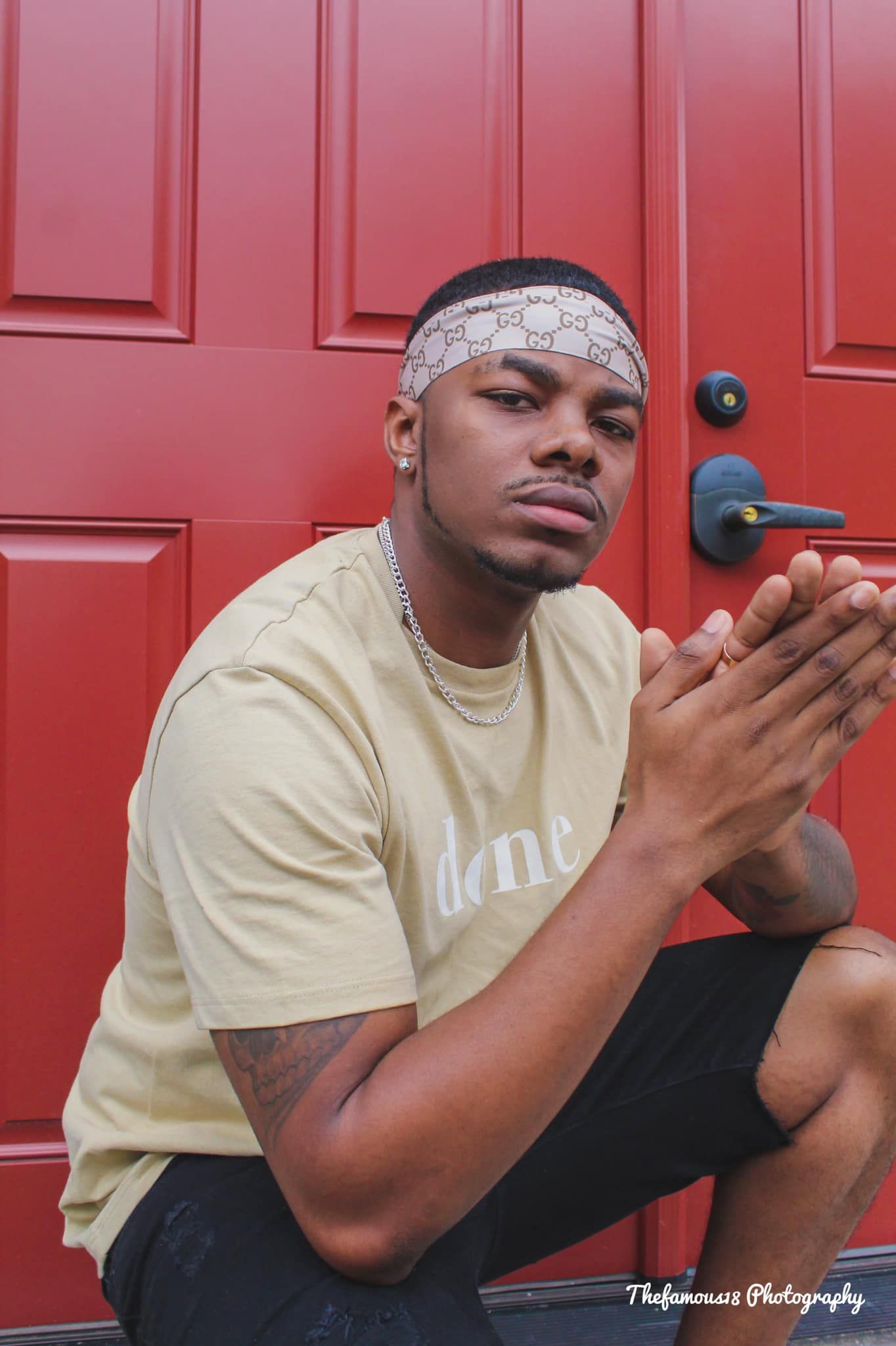 Young man in yellow tee with Gucci headband sits against red door.