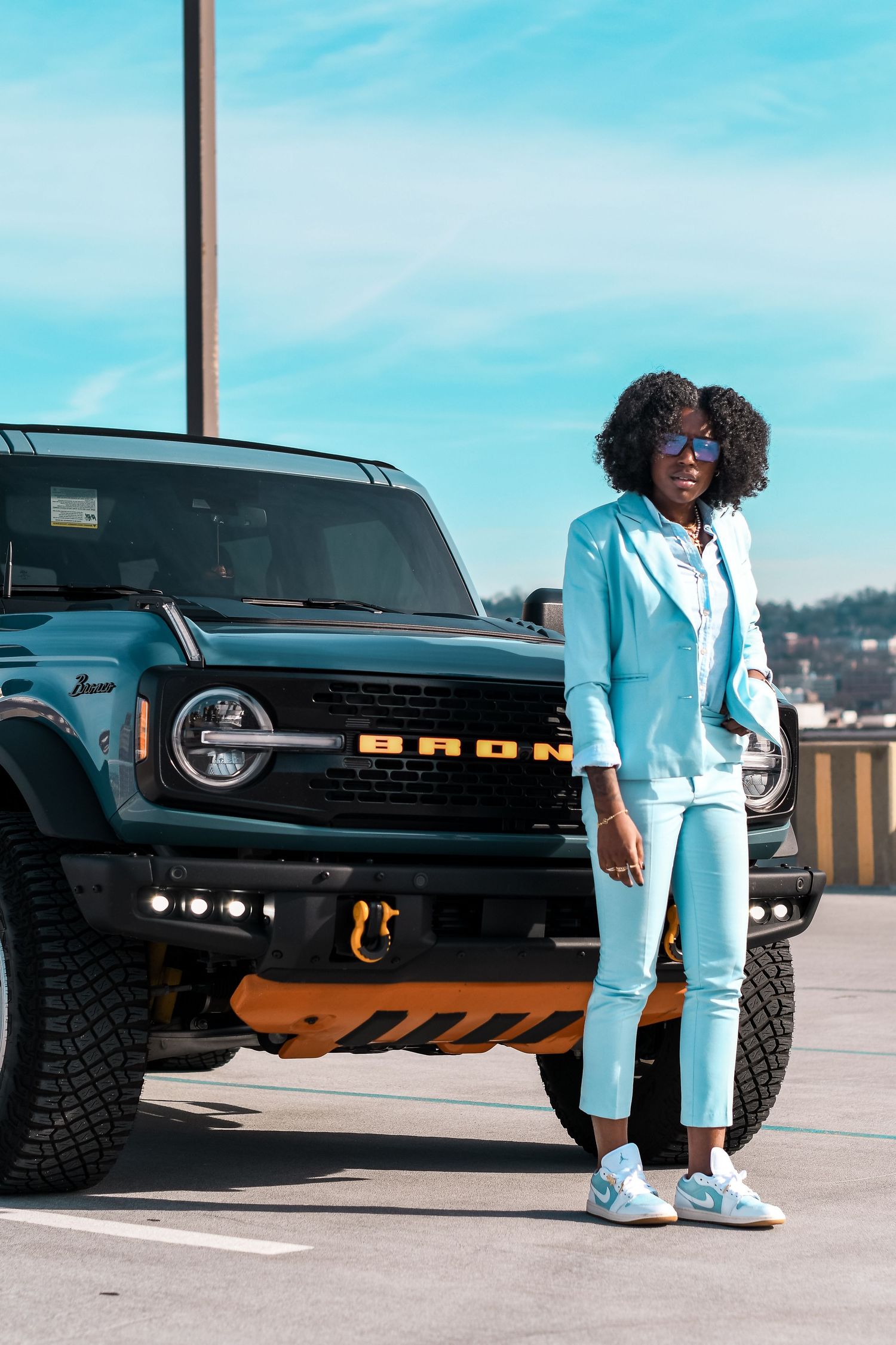Black woman in light blue suit posing confidently in front of a teal Ford Bronco outdoors.