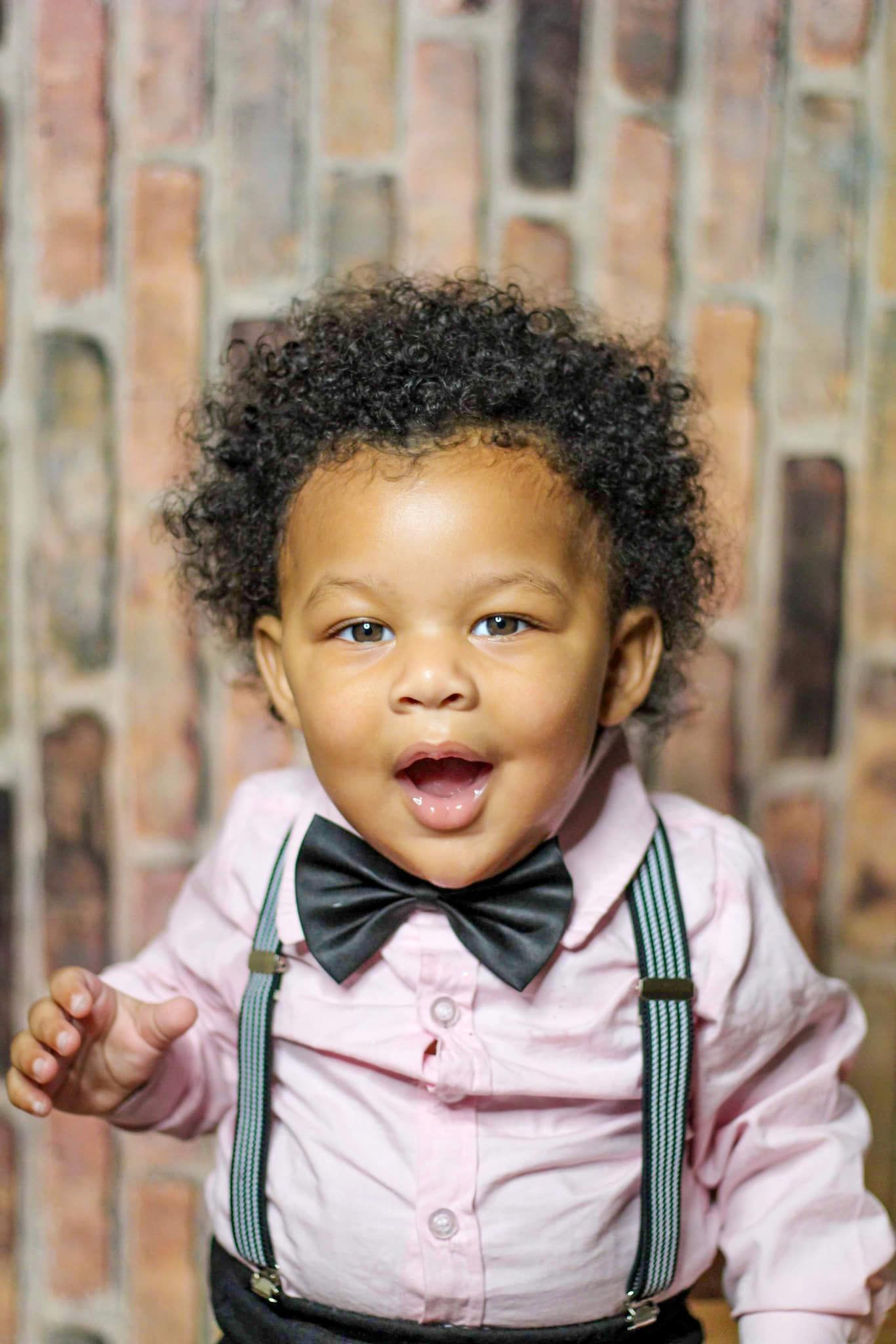 Adorable curly-haired toddler in pink shirt with bow tie and suspenders against brick wall.