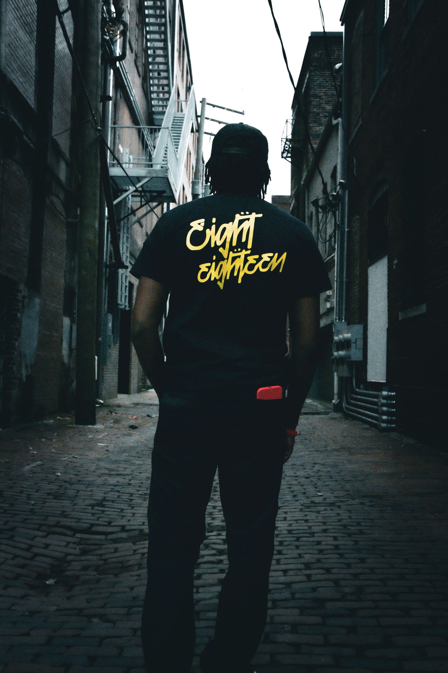 Man in black Eight Eighteen t-shirt stands in dark urban alleyway.
