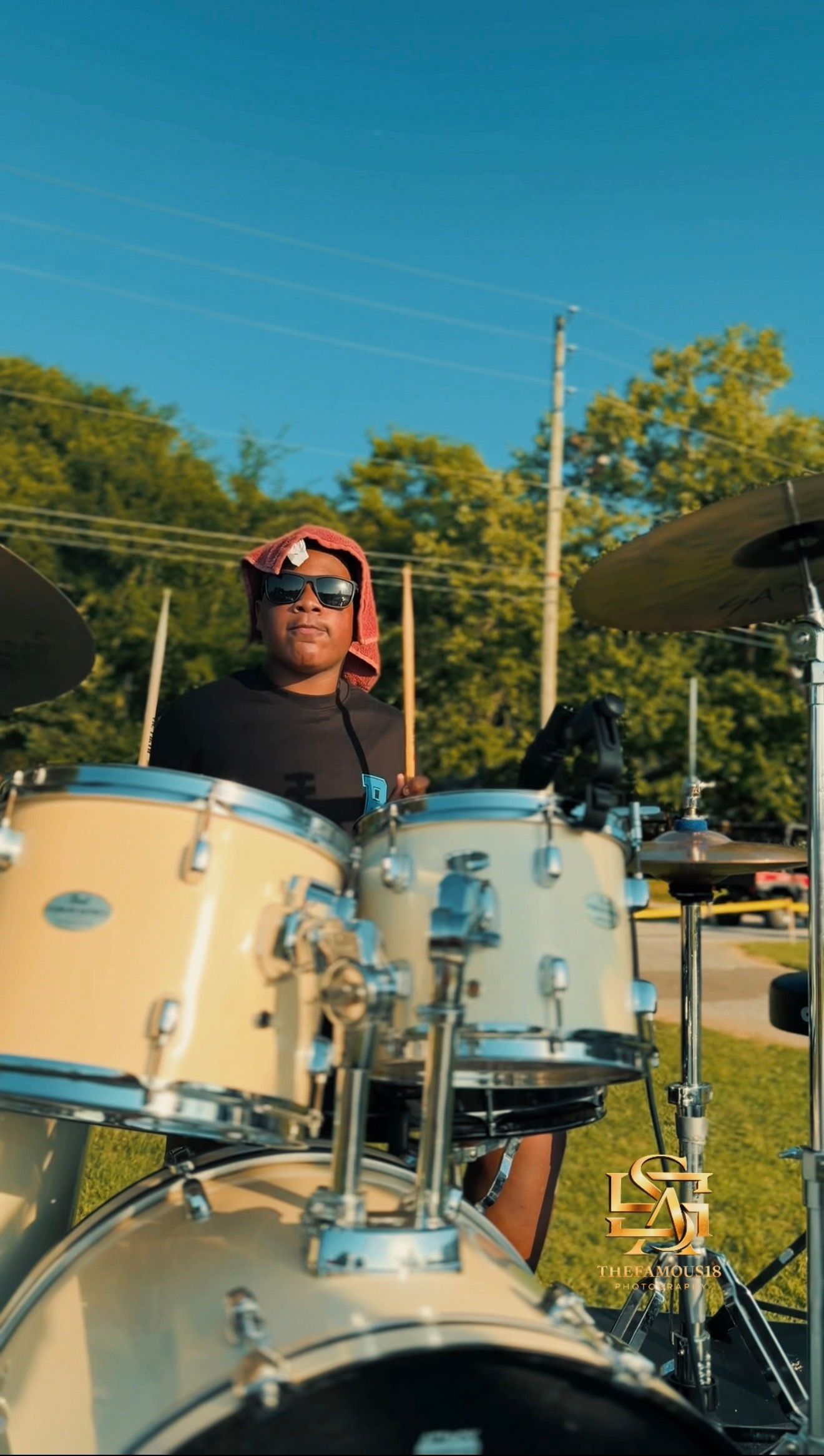 A drummer plays a white drum kit outdoors under a clear blue sky, wearing sunglasses and a red hoodie.