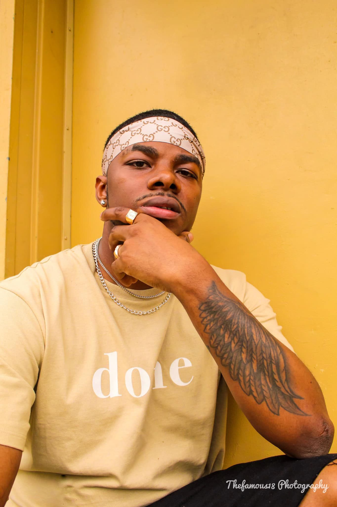 A young Black man in a beige done t-shirt and Gucci headband poses thoughtfully against a yellow wall.