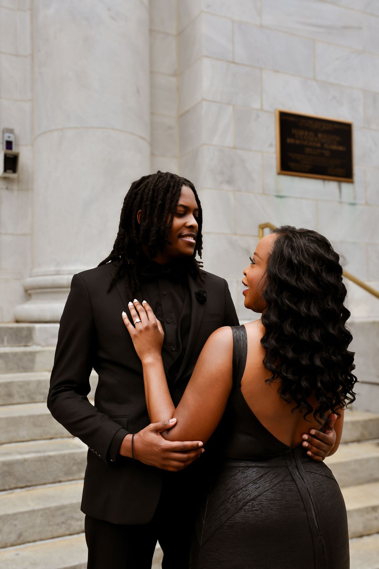 Smiling couple in black formal attire embracing on stone steps outside a grand building.