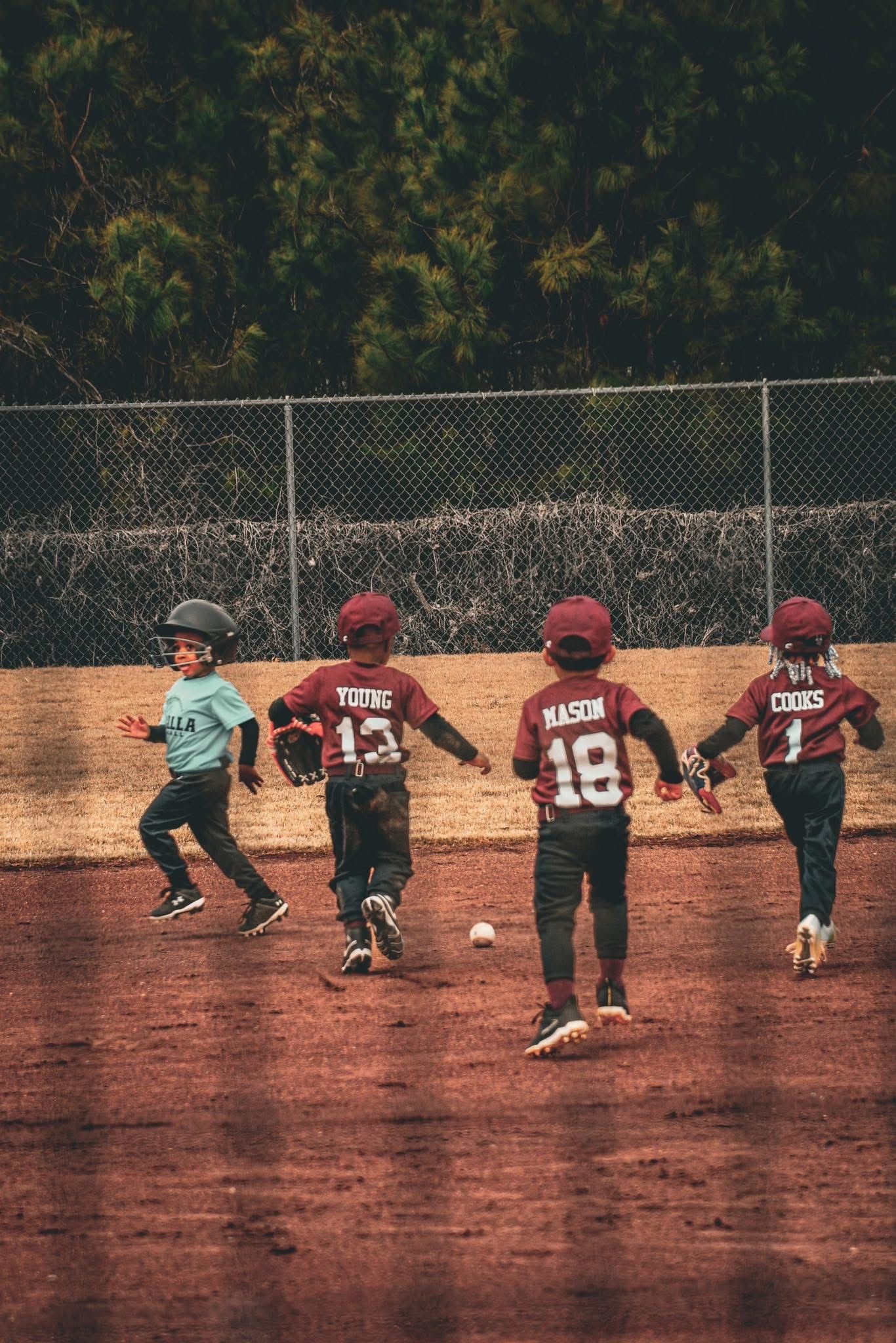 Young youth baseball players in maroon uniforms chasing a ground ball on a dirt infield during a game.