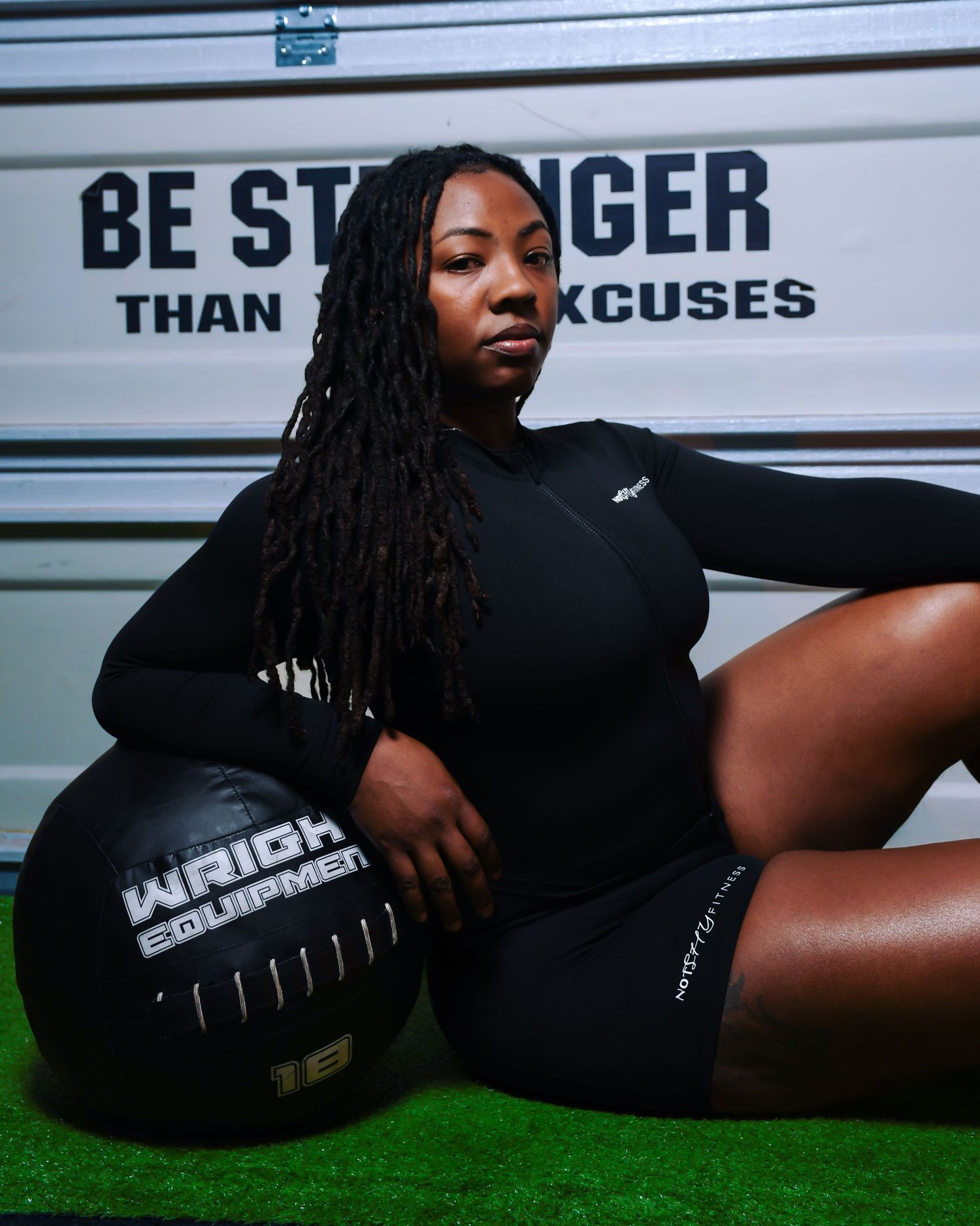 Black woman with locs in black workout outfit leaning on medicine ball in gym.