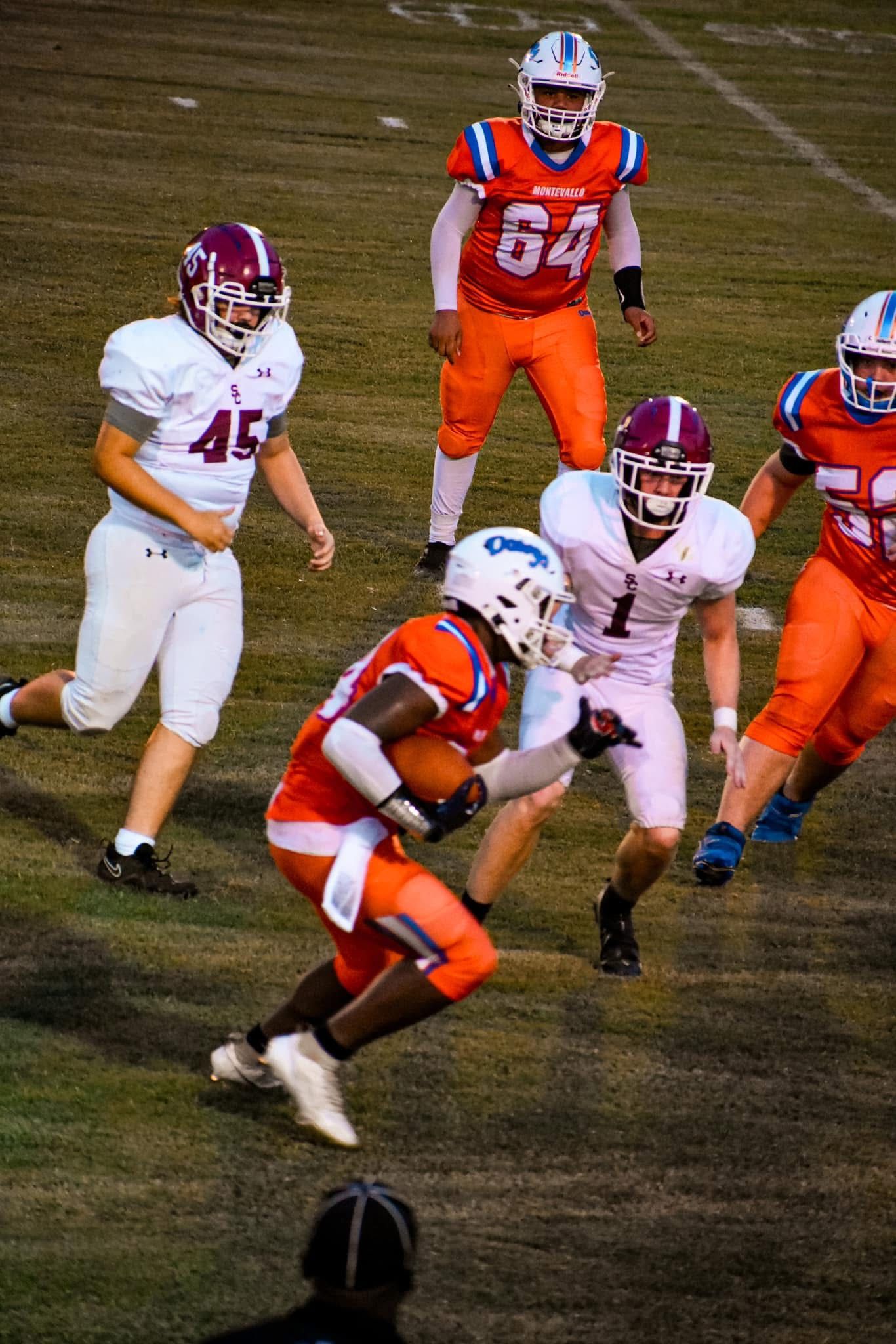 High school football players in white and maroon uniforms run a play against an orange-uniformed team at night.