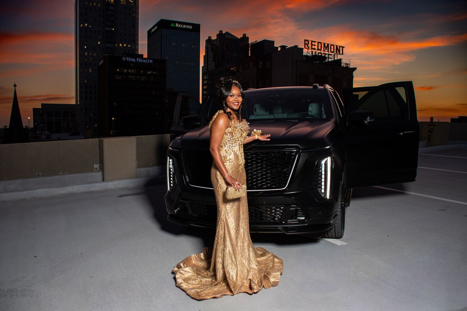 Woman in gold mermaid gown poses beside black luxury SUV at colorful urban sunset.