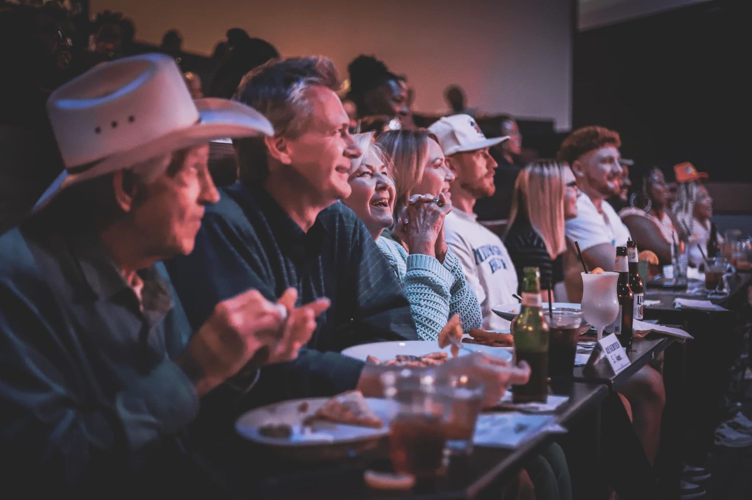 Diverse audience laughing and clapping at a live event with food and drinks on tables.