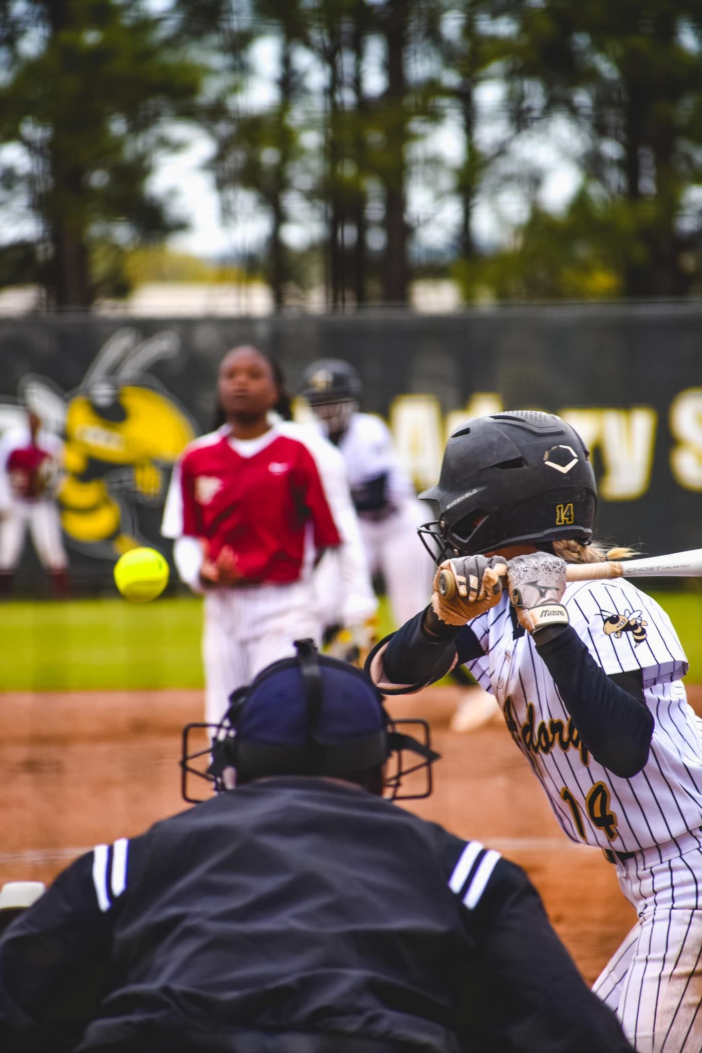 A softball batter in a gold and black uniform swings at a pitch during a competitive game.