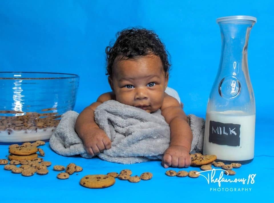 Adorable baby lying on grey blanket surrounded by cookies and milk bottle on blue background.