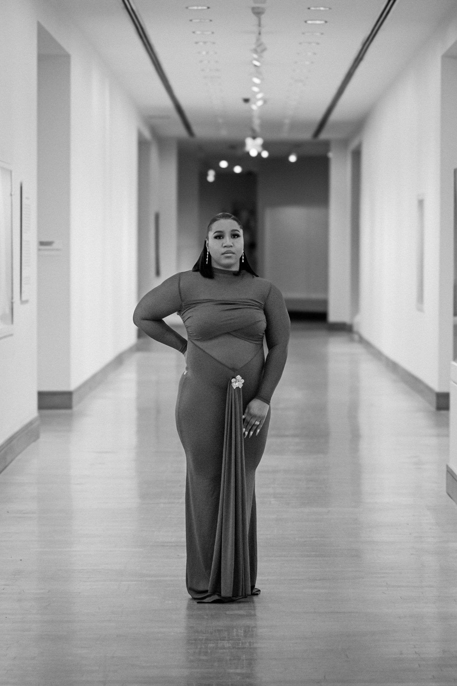 Elegant woman in form-fitting gown stands in black and white art gallery hallway.