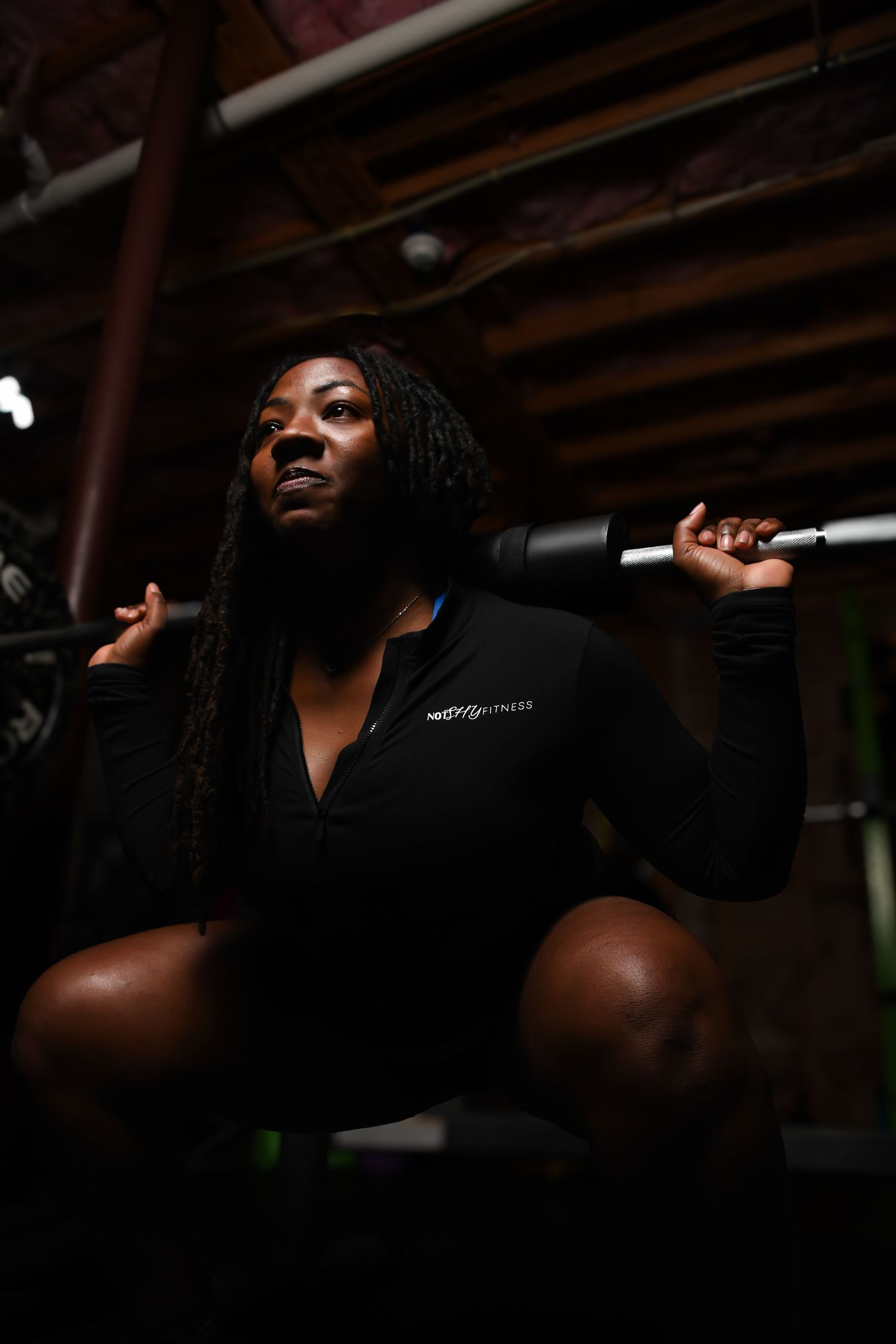 A determined woman performs a barbell squat in a dark, rustic gym wearing a black fitness long-sleeve top.