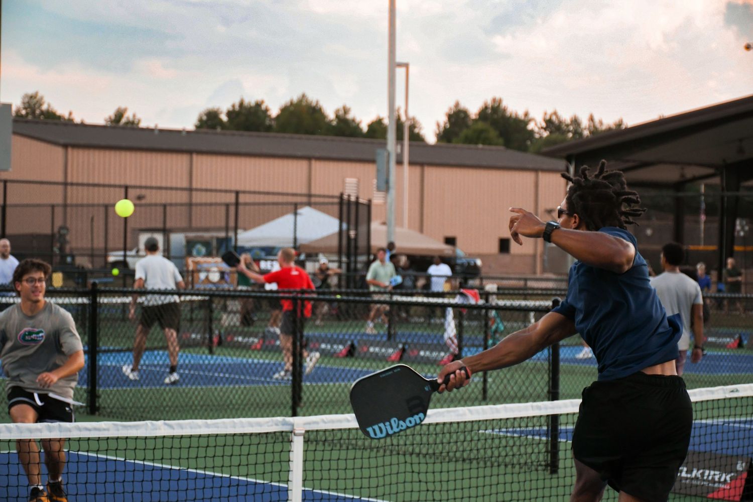 Players compete in an outdoor pickleball tournament on blue courts, with spectators watching under evening lights.