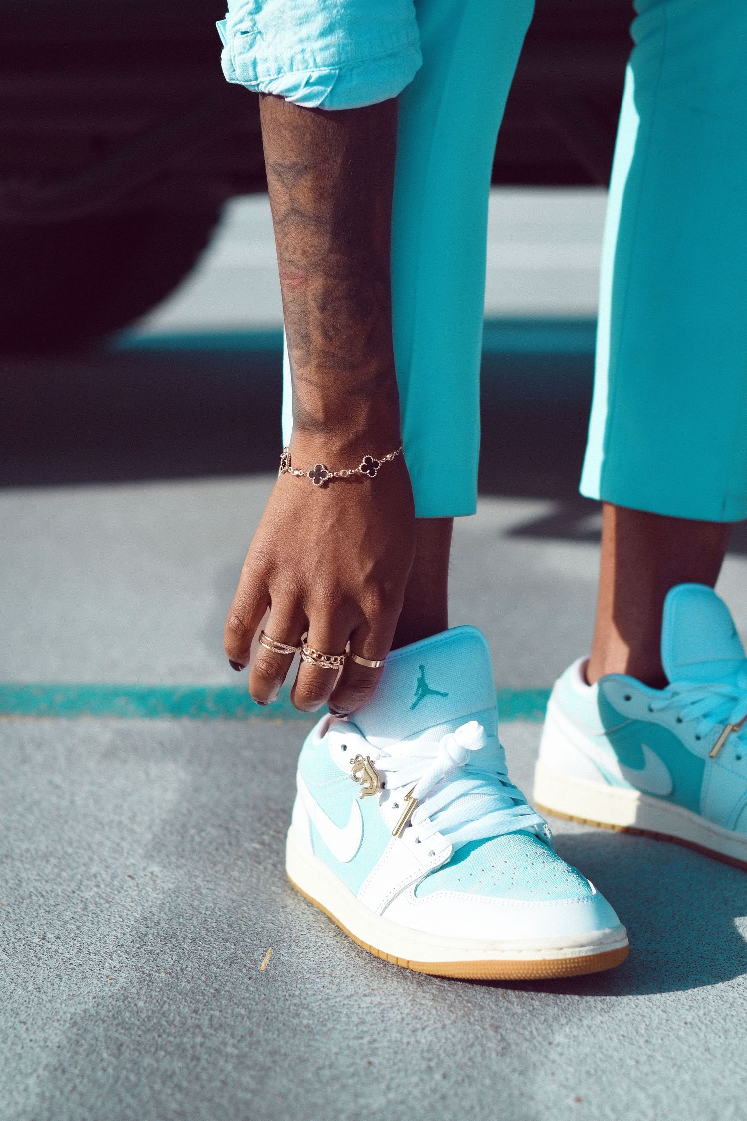 Person wearing teal outfit and white Air Jordan 1 Low sneakers with gold jewelry and tattoos on hand.