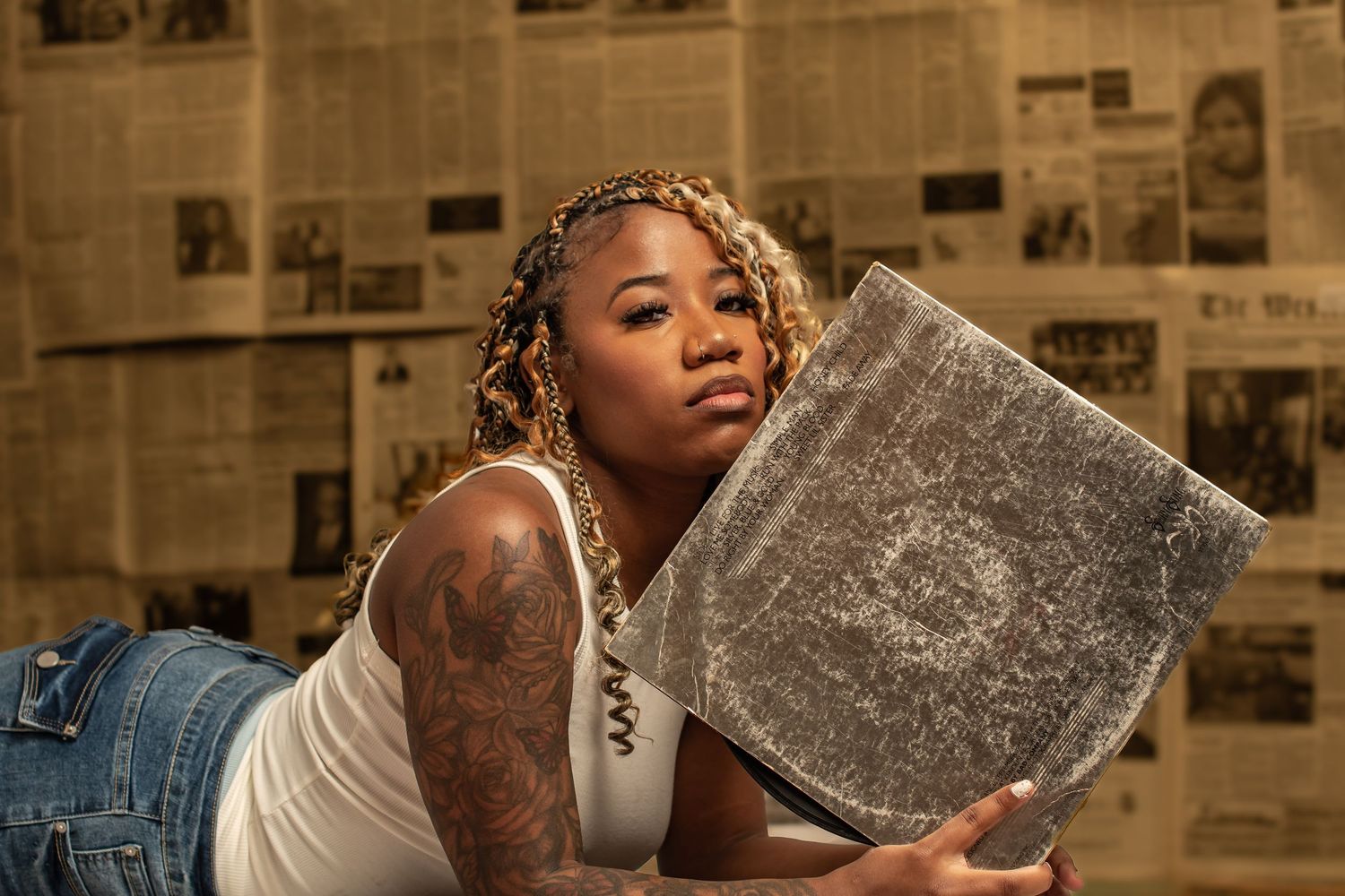 Tattooed woman with braids poses with a vinyl record against a newspaper-covered wall background.