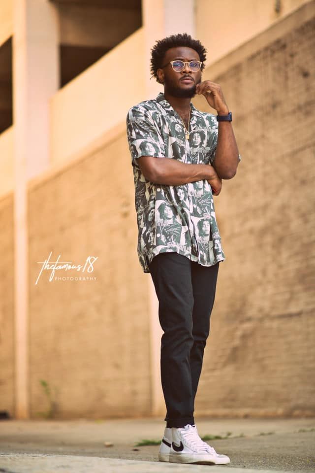 A fashionable Black man in a printed shirt and glasses stands confidently in an urban outdoor setting.