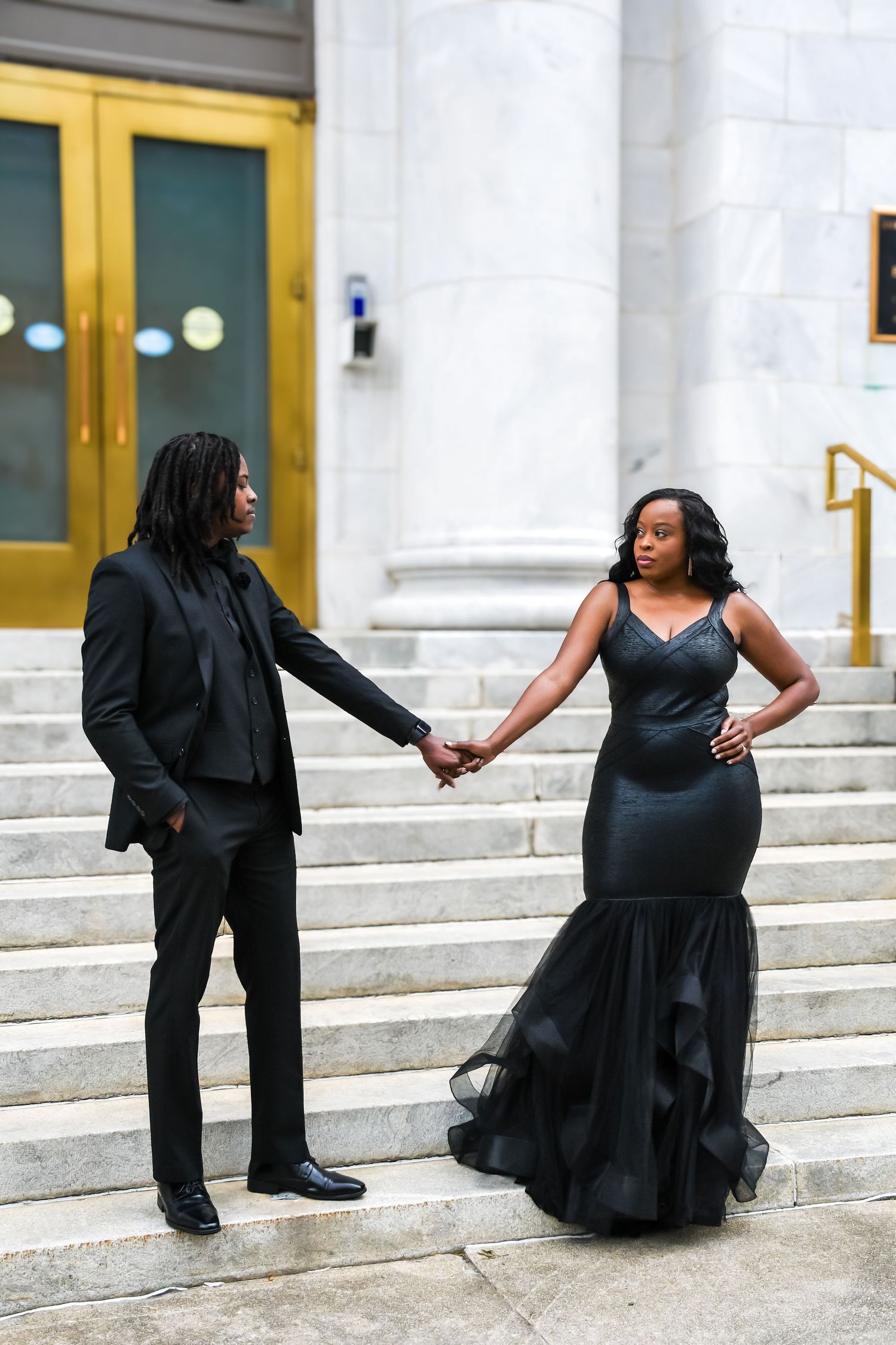 Couple in matching black formal attire holding hands on marble steps of a grand building.