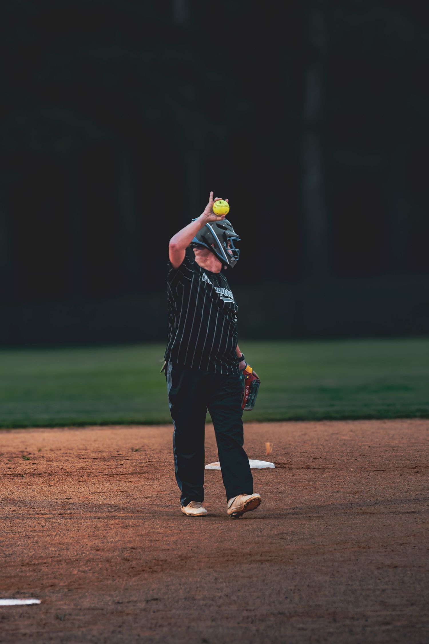 Softball player in black pinstripe uniform catching a yellow ball overhead on a dirt infield at dusk.