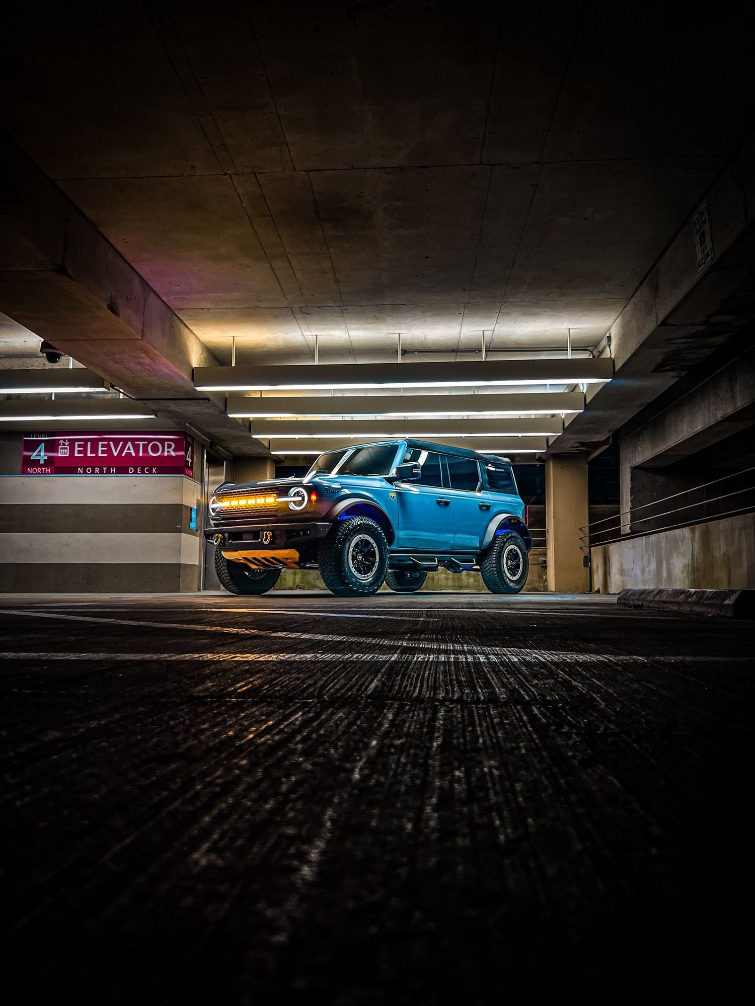Blue Ford Bronco parked dramatically in a dimly lit parking garage near an elevator sign.