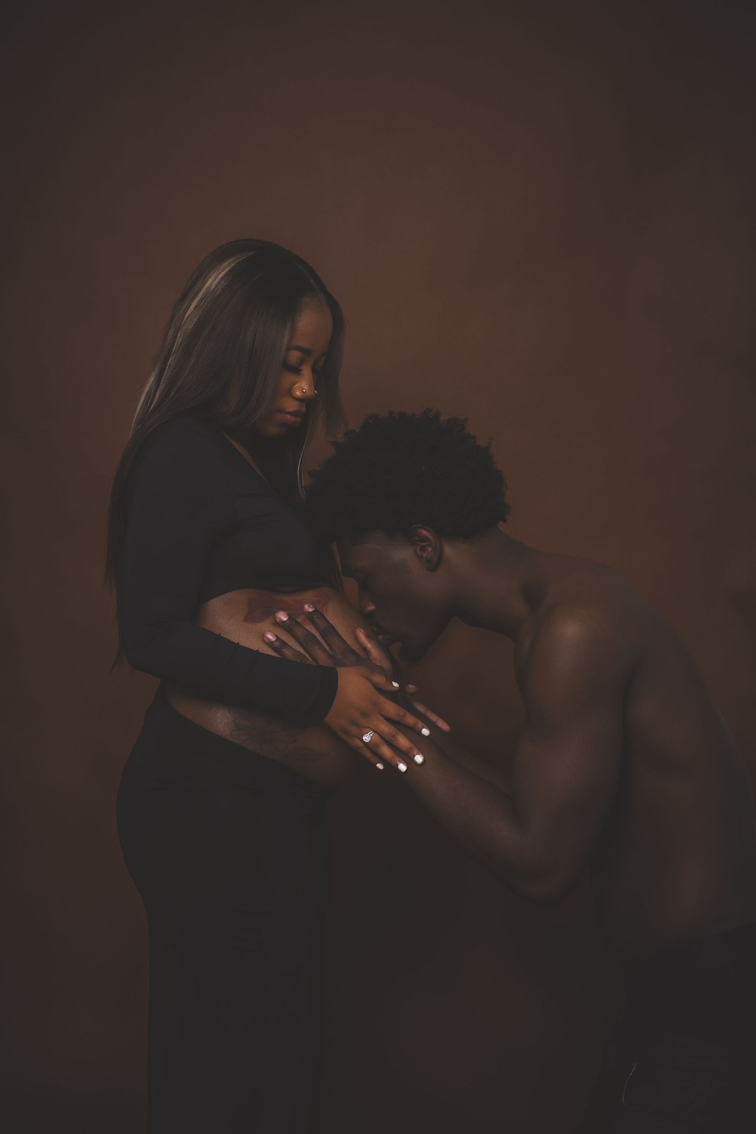 An expecting couple poses tenderly in a dark studio, the man lovingly pressing his face to the woman's pregnant belly.
