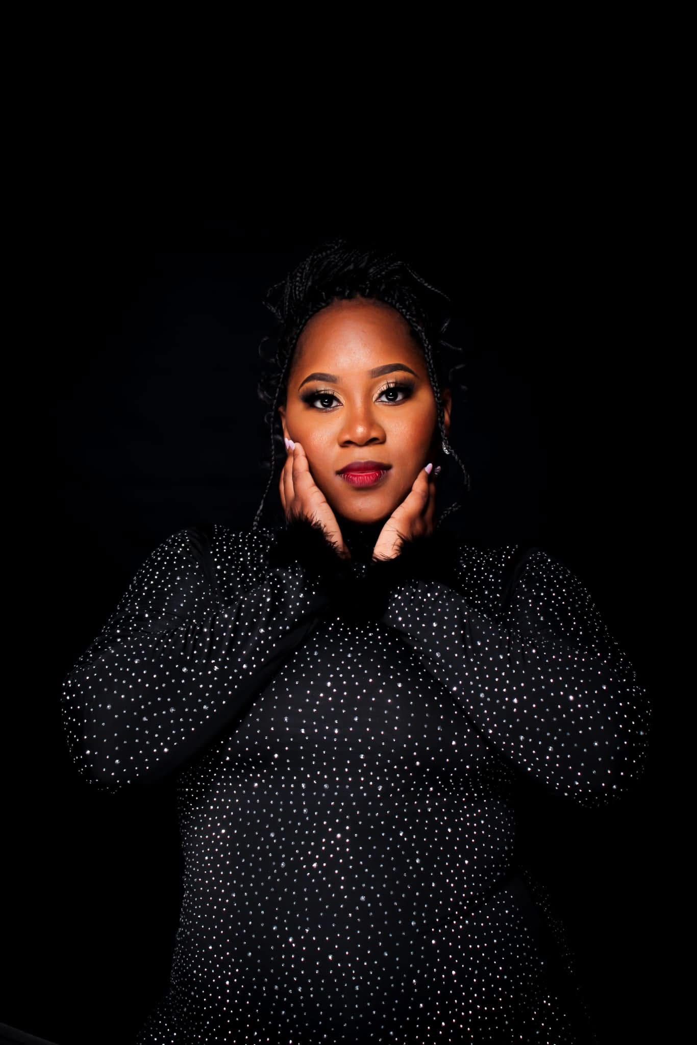 Woman in black sparkle top posing with hands on cheeks against dark background.
