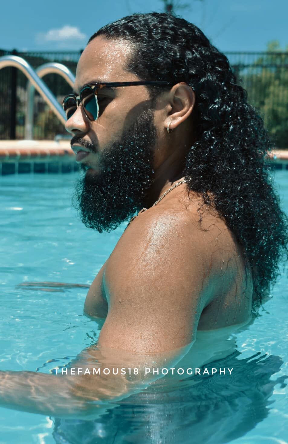 Man with long curly hair and beard wearing sunglasses stands in a swimming pool on a sunny day.