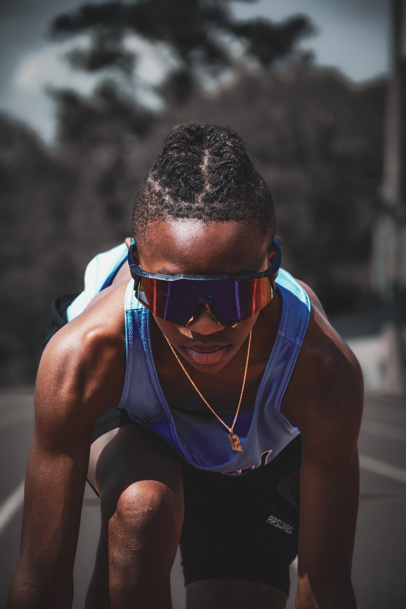 A focused athlete in a blue track jersey and mirrored sunglasses crouches at the starting line.