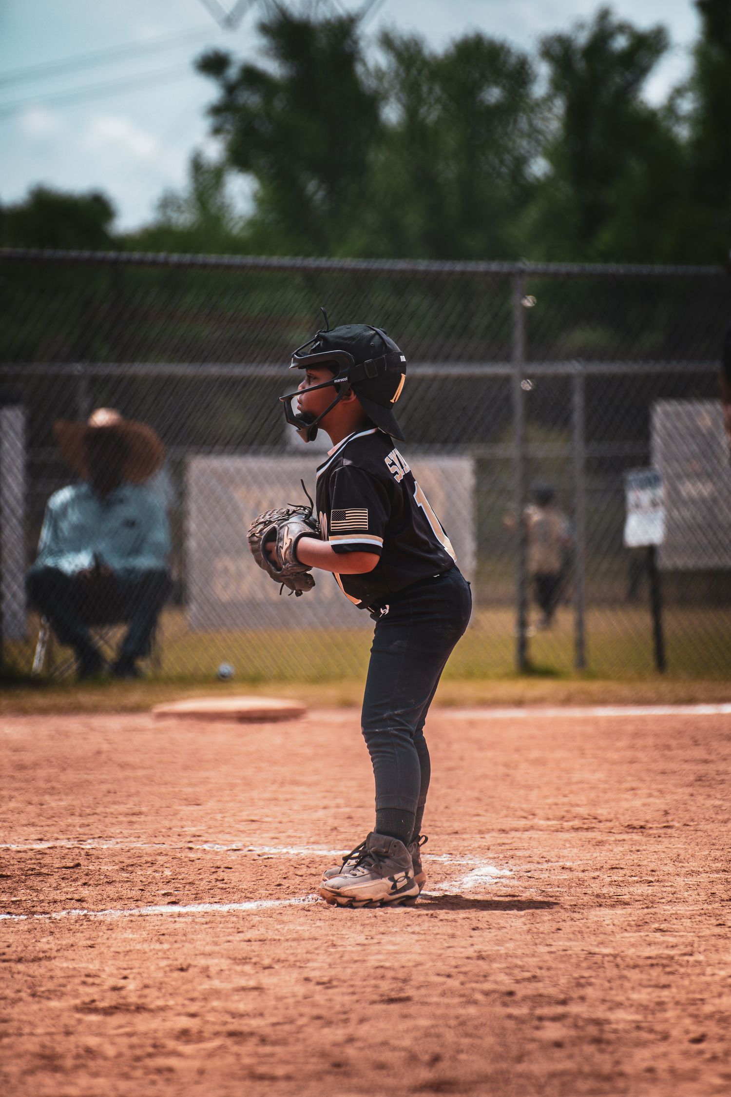 Young baseball player in black uniform stands ready at home plate on a dirt field during a youth league game.