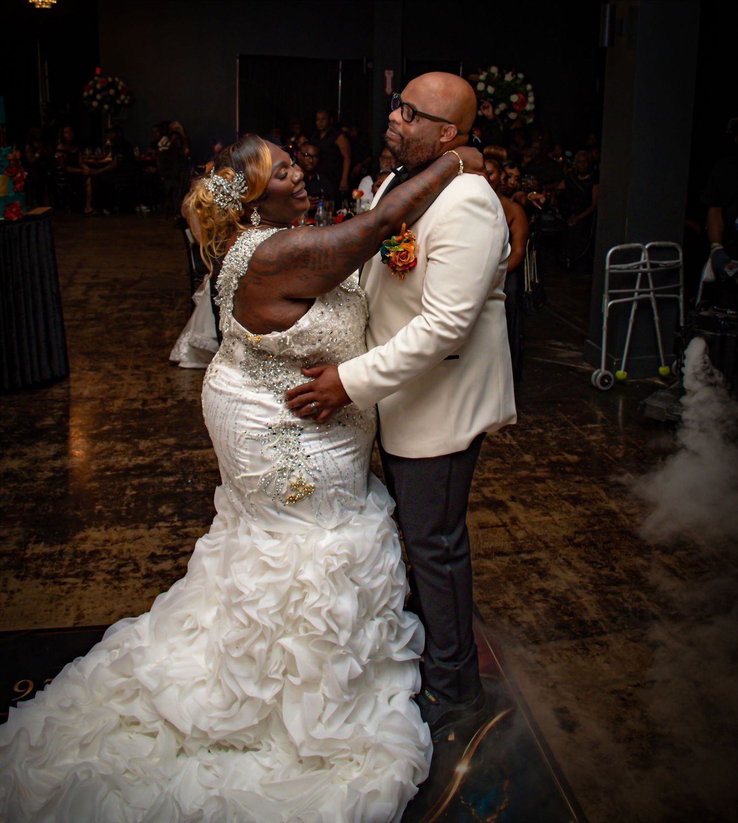 Bride and groom share first dance at wedding reception, bride in ruffled white gown, groom in ivory suit.