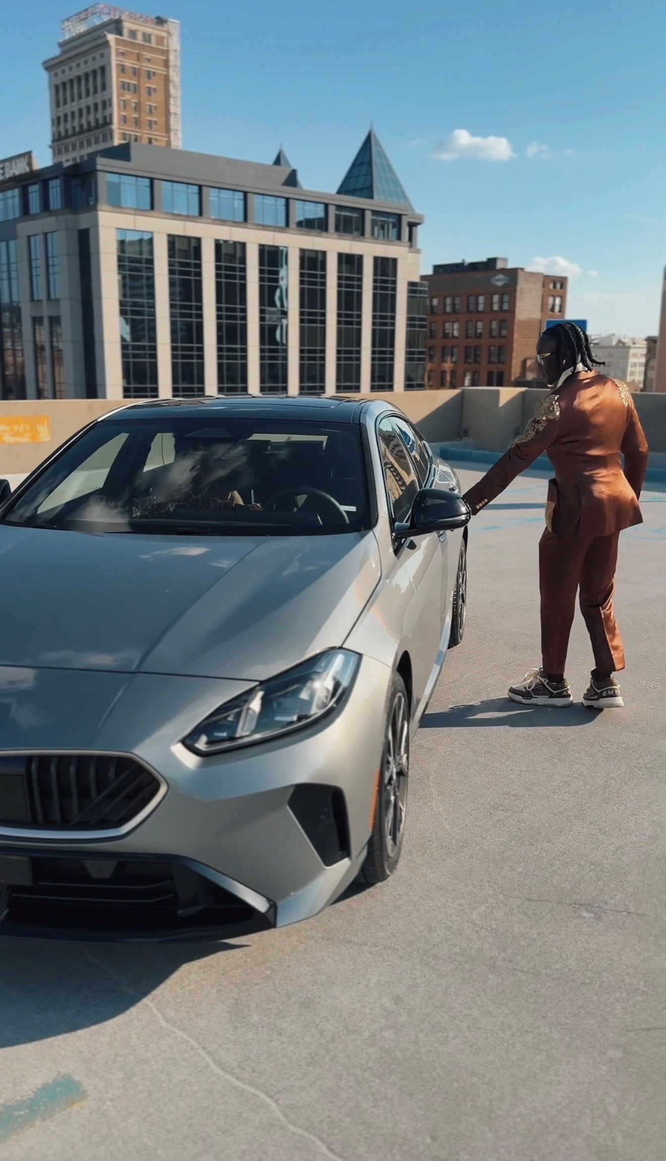 Person in rust-colored outfit standing beside a silver sedan on a rooftop parking lot with city buildings in background.