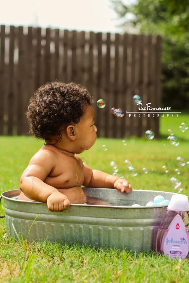 An adorable baby sits in a vintage metal tub outdoors watching colorful soap bubbles float by.