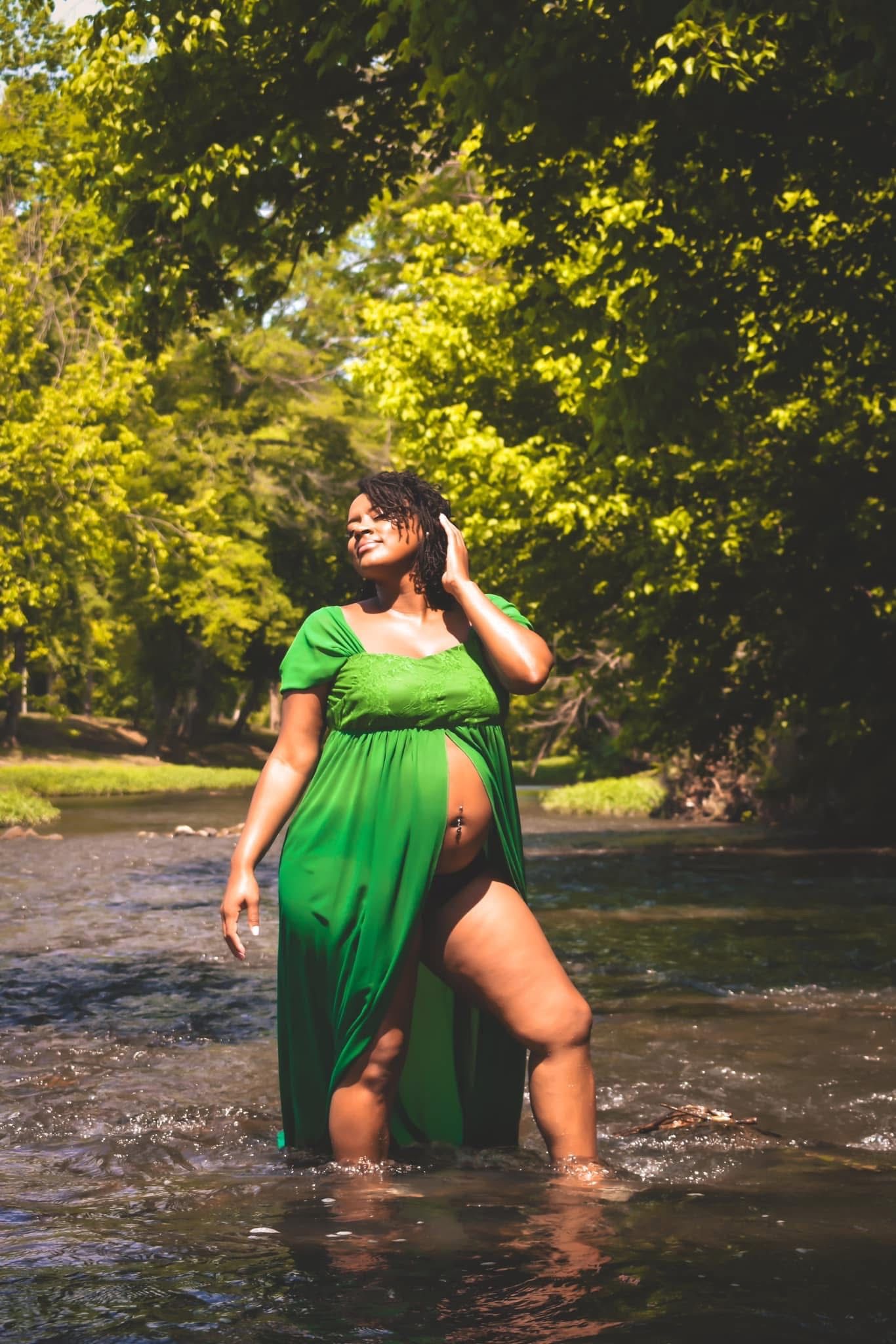Pregnant Black woman in flowing green dress standing in shallow river surrounded by lush trees.
