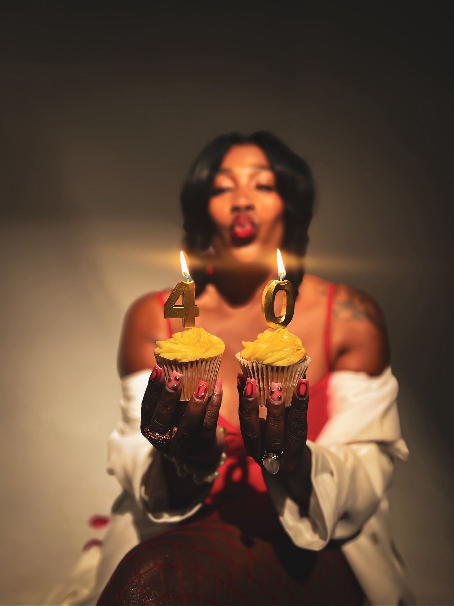 Woman blowing out gold number 40 birthday candles on cupcakes in a dramatic low-light setting.