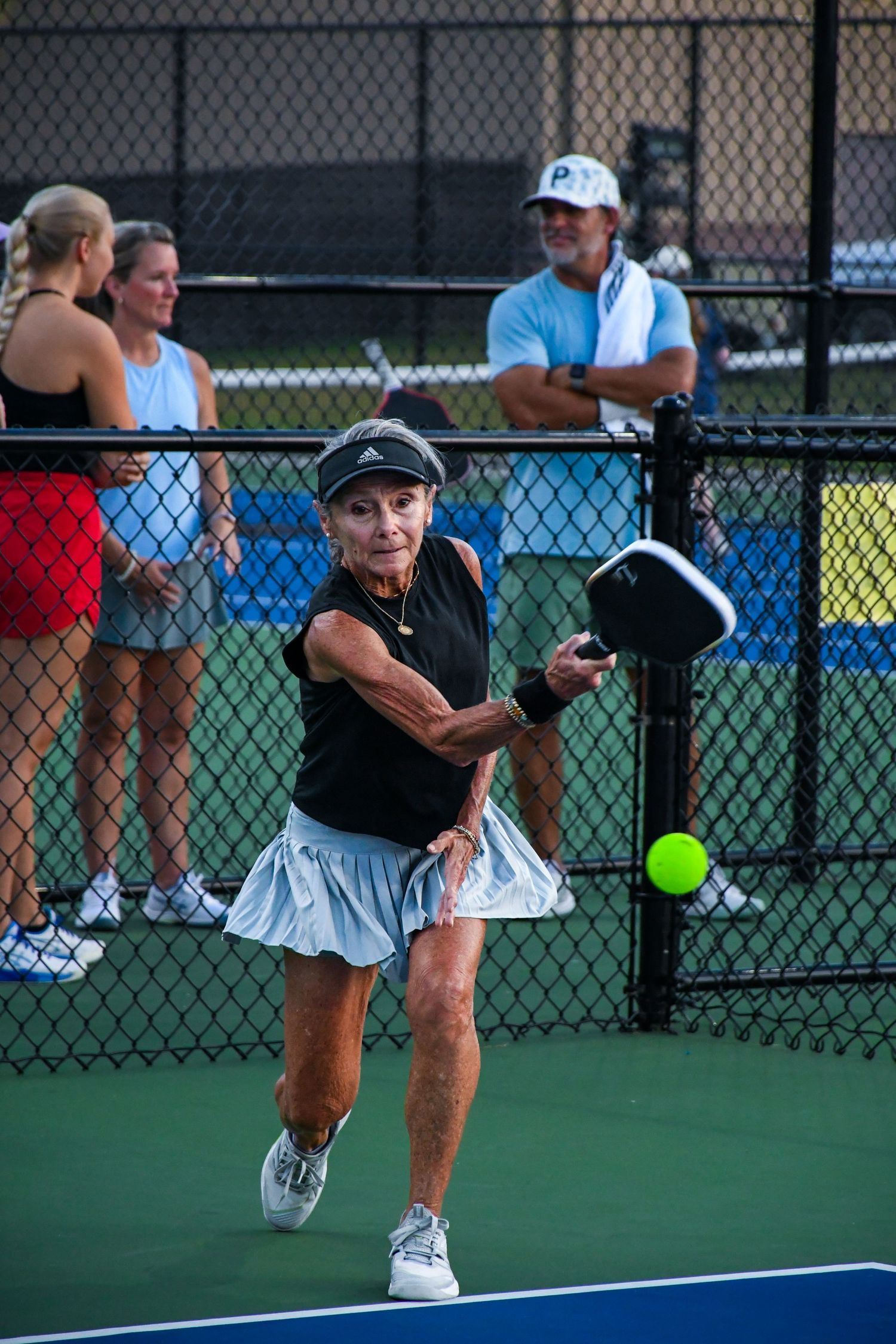 Senior woman playing pickleball, hitting ball with paddle on outdoor court while spectators watch in background.