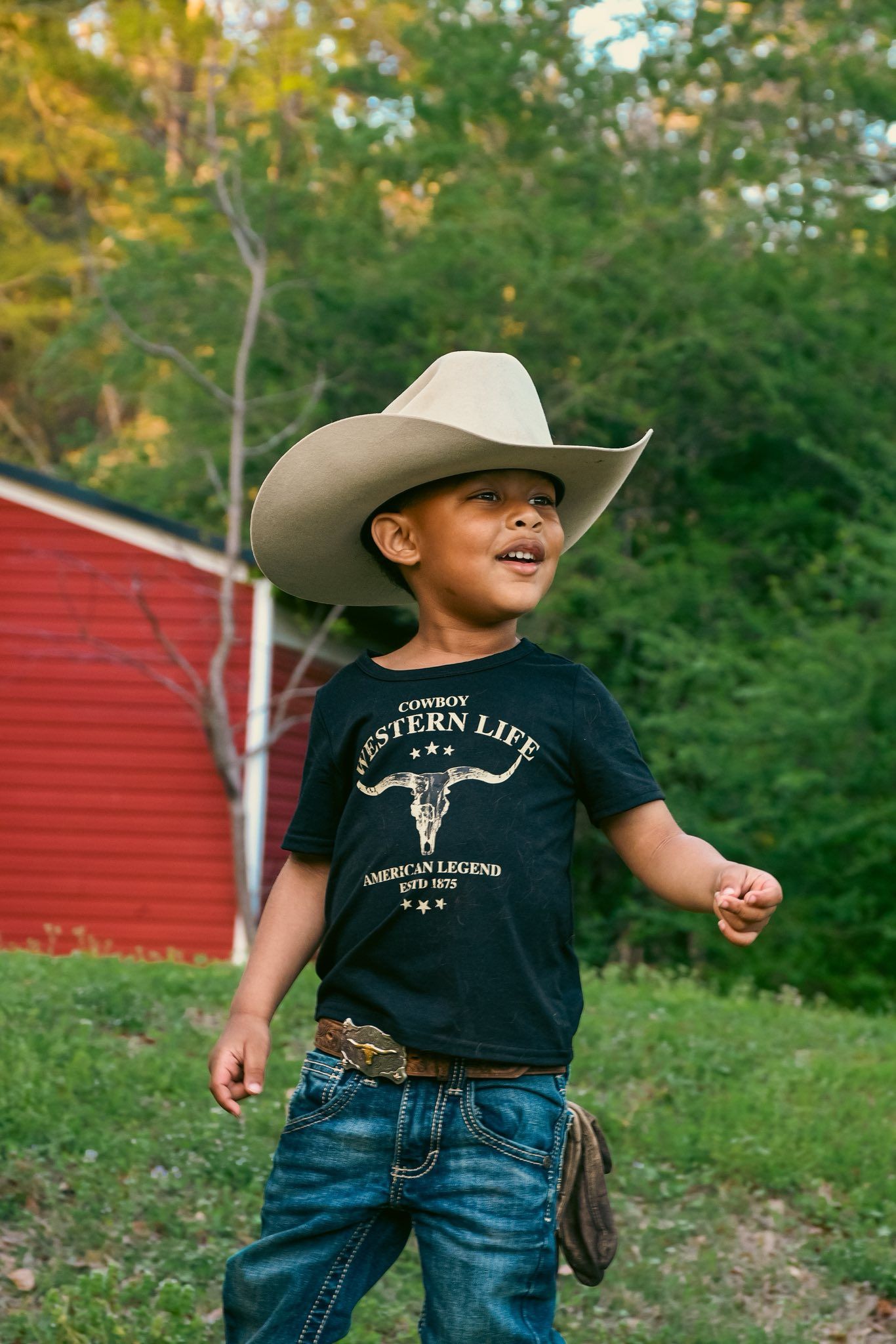 Young boy in cowboy hat and Western Life t-shirt walks outdoors near red barn.