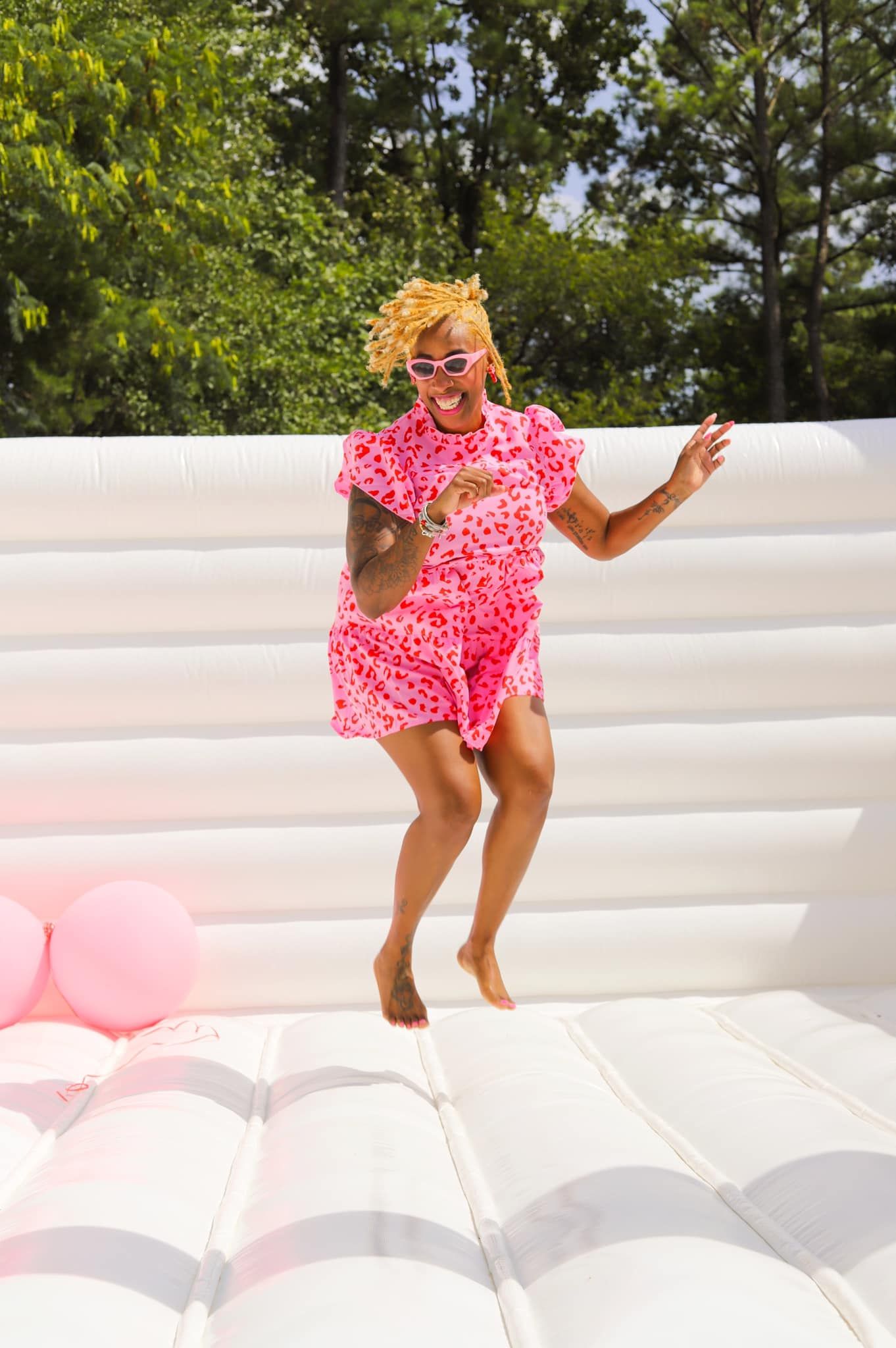 Joyful woman in pink floral outfit jumping on white inflatable bounce house.