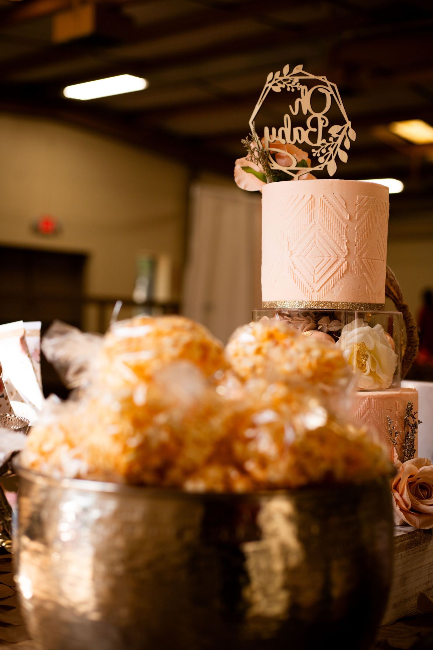 An elegant single-tier pink wedding cake with a gold topper displayed behind a bowl of golden treats.