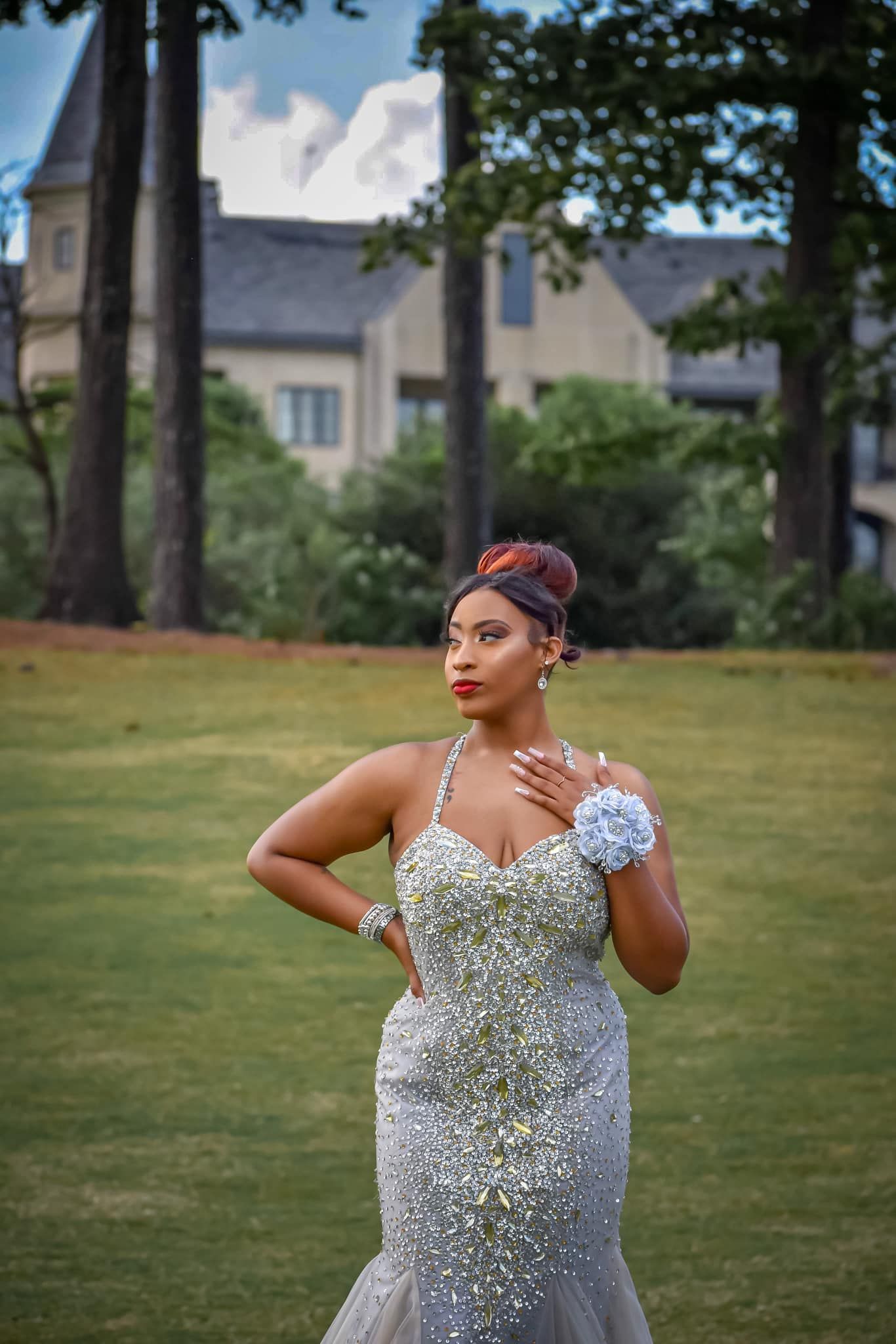 A woman in a silver beaded mermaid gown poses gracefully on a green lawn near a mansion.