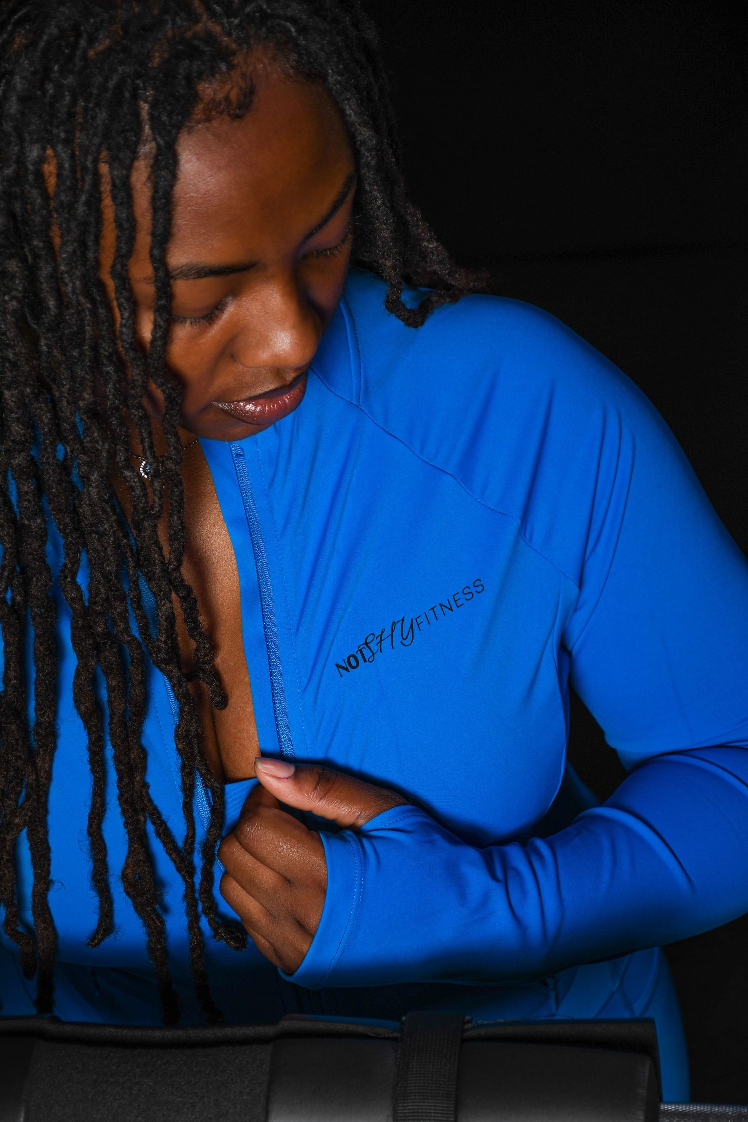 Woman with locs wearing a blue athletic zip-up jacket with fitness branding, looking down in a dark studio setting.