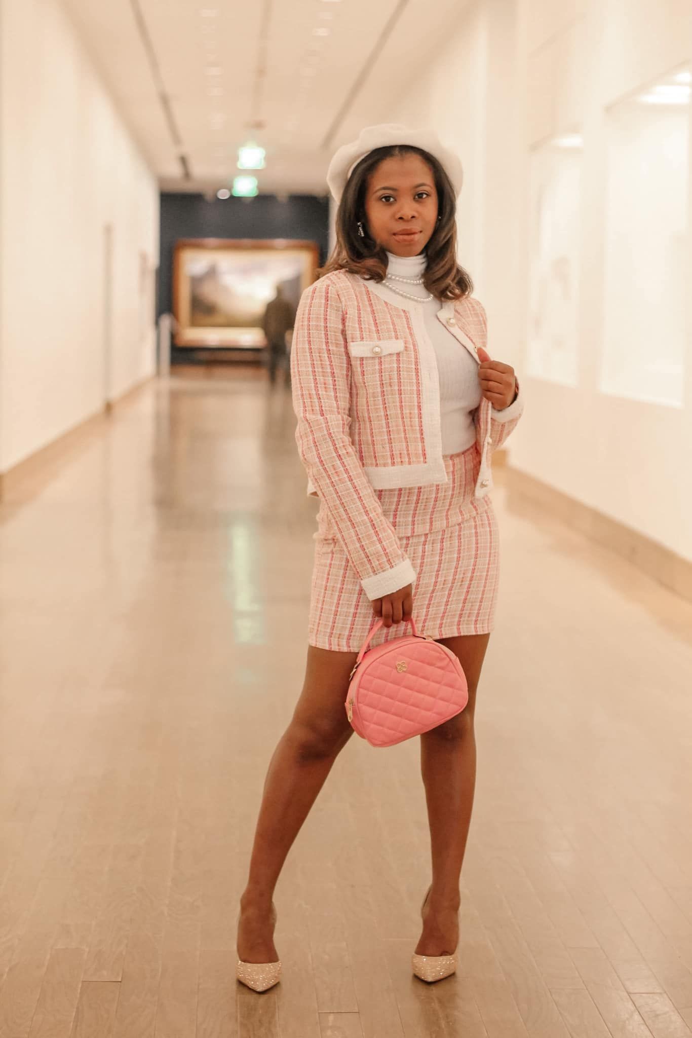 A chic Black woman in a pink plaid skirt suit and beret poses in an elegant museum hallway.