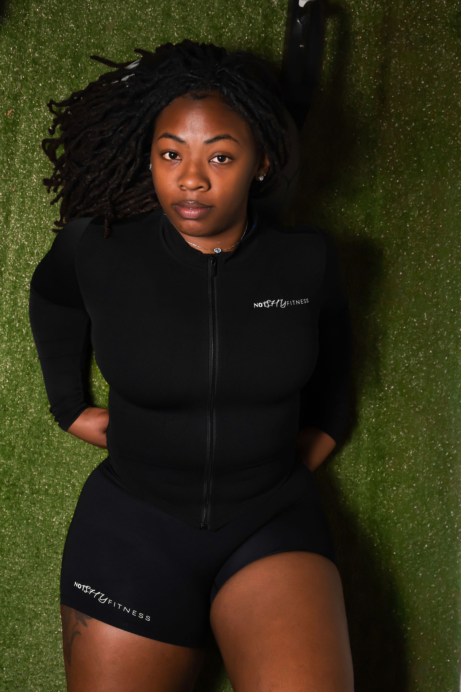 Woman in black NorthFit fitness jumpsuit lying on green turf grass looking at camera.