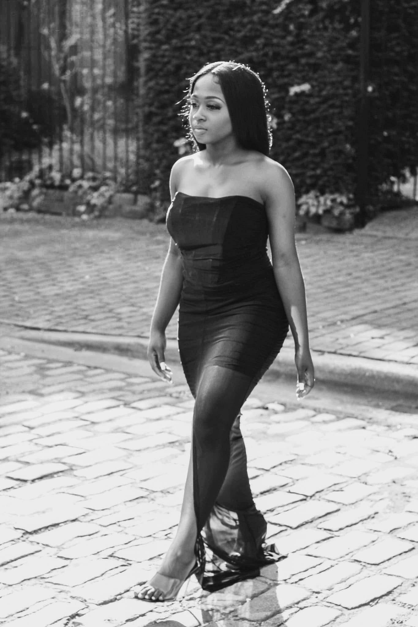 A woman in a strapless black gown walks on a cobblestone street in black and white photo.
