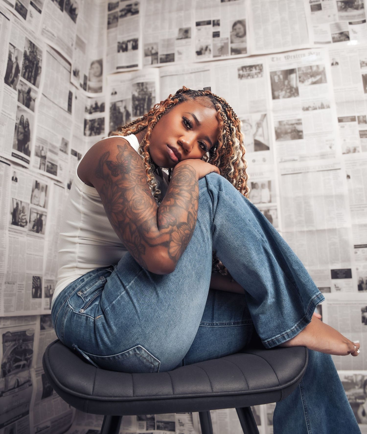 Tattooed woman with braids sits on a stool against a newspaper wall backdrop in jeans and white top.