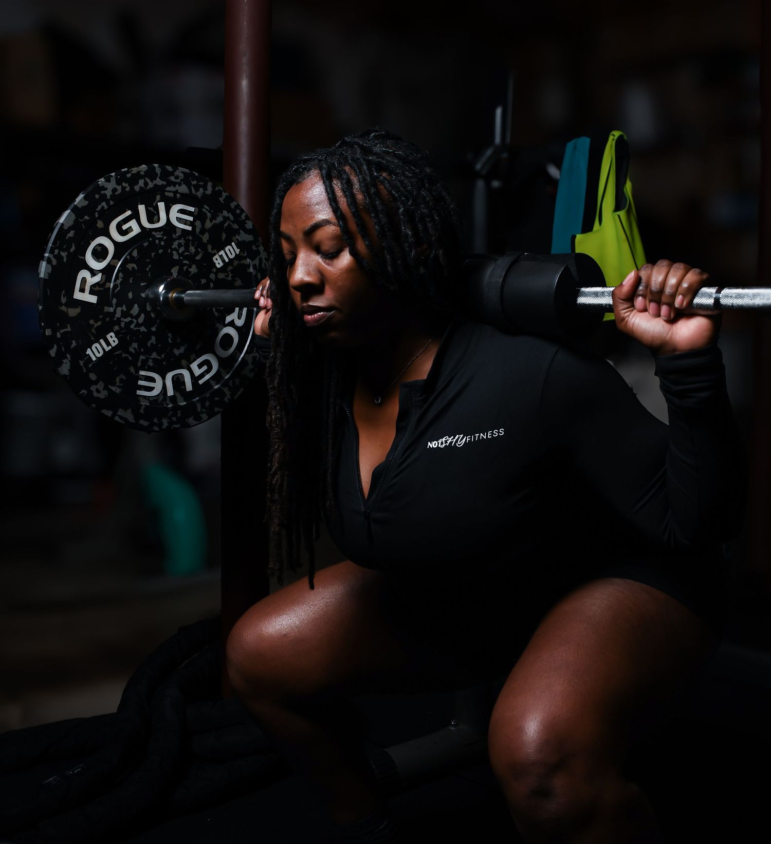 A Black woman athlete squats with a Rogue barbell on her shoulders during a weightlifting session in a dark gym.