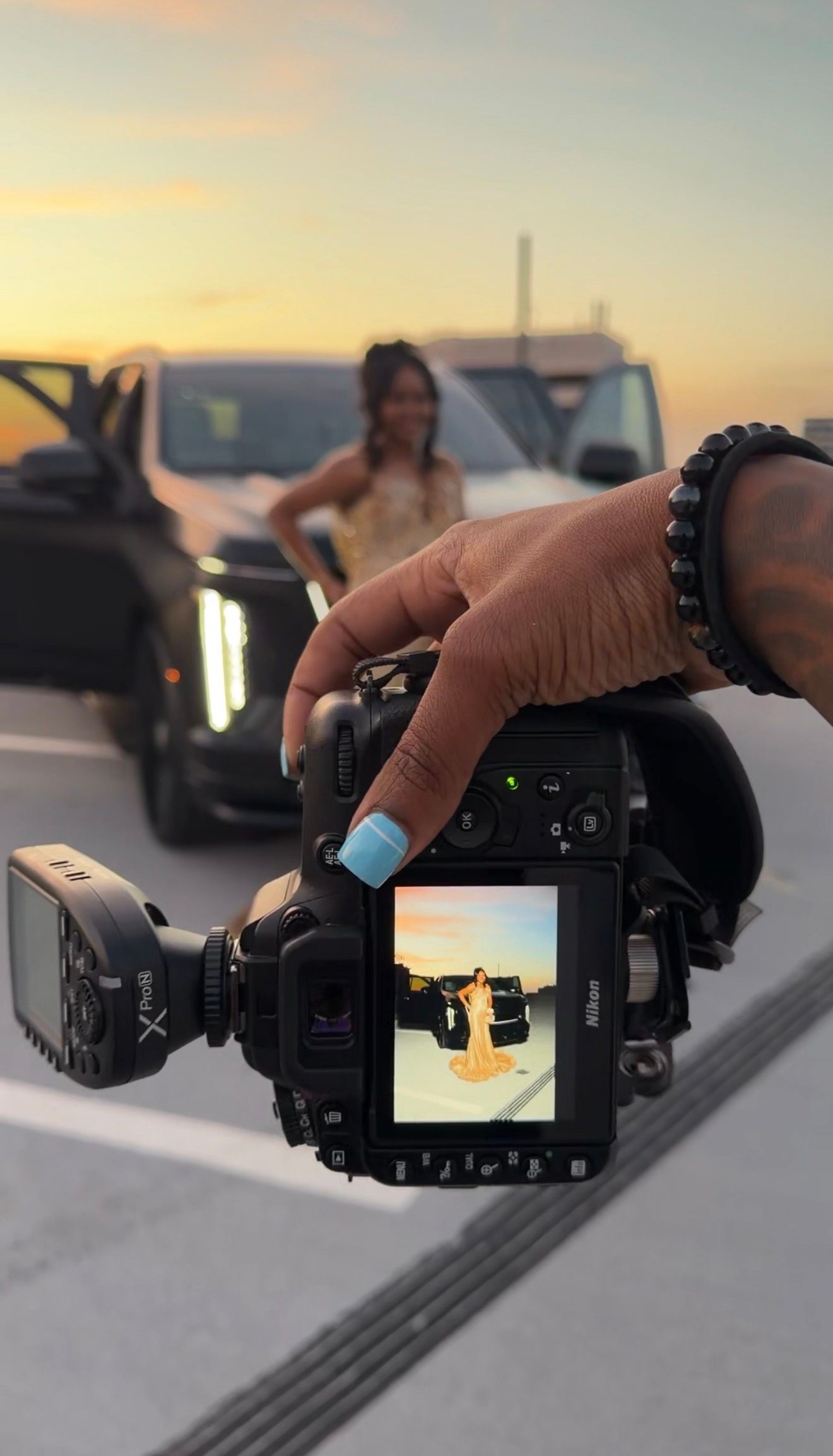 Photographer holds Nikon camera displaying woman in yellow dress on screen during golden hour photoshoot near vehicles.