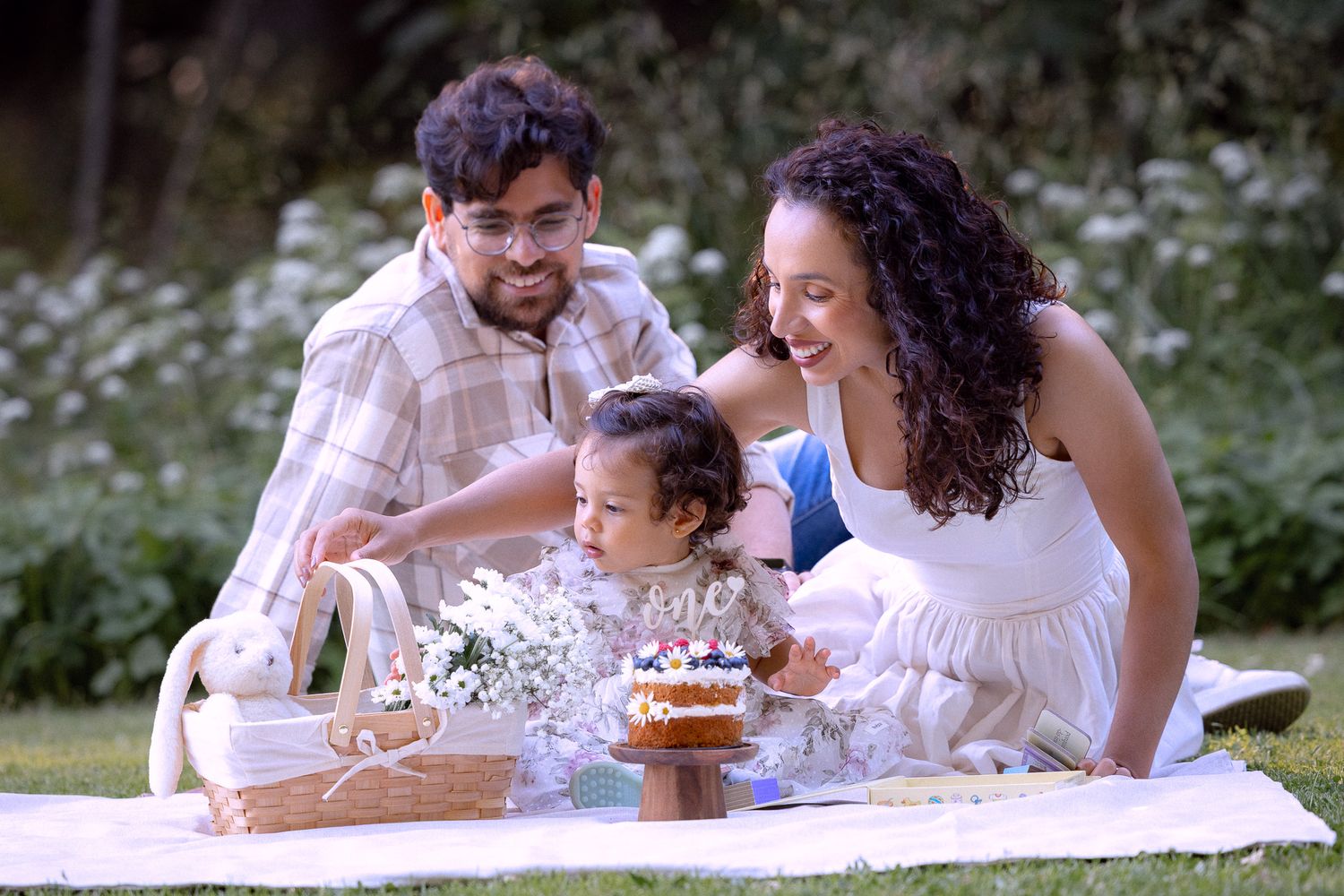Parents celebrating toddler's first birthday with cake on picnic blanket surrounded by flowers in a garden.