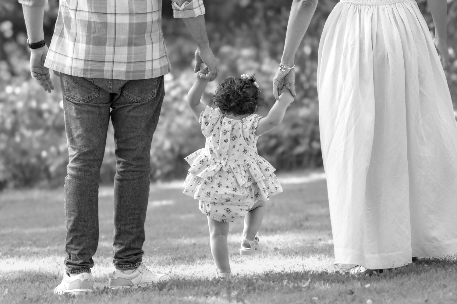 Black and white photo of parents holding toddler girl's hands as she walks in a garden.